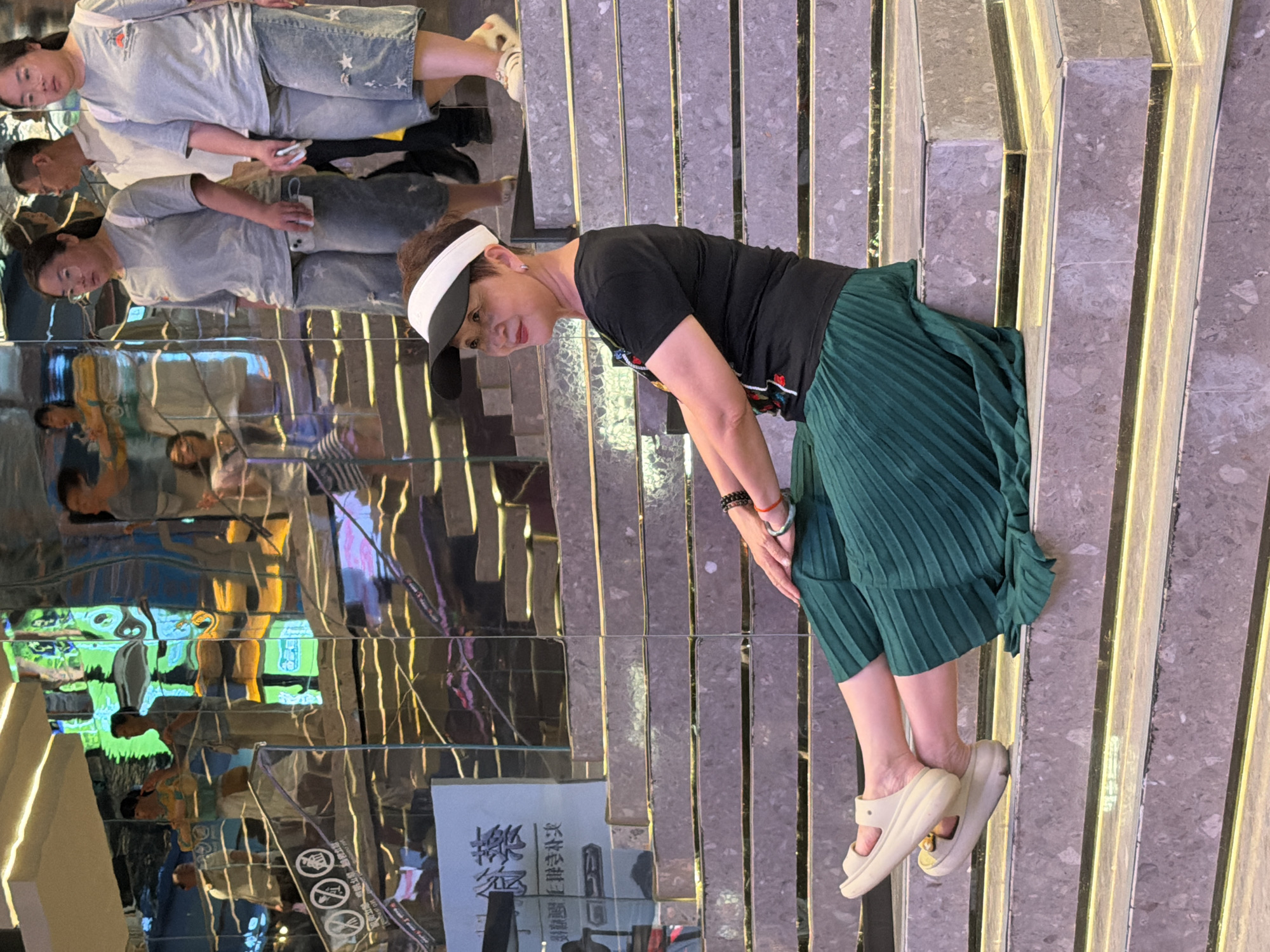 A middle-aged East Asian woman is sitting on marble stairs with her legs extended, wearing a black t-shirt, a green pleated skirt, and cream-colored slides. She has a black and white visor on and is looking to her left. Her hands are adorned with bracelets and a ring. In the background, on a higher level of the stairs, two other women are walking, one wearing a light grey t-shirt and denim shorts with white stars, and the other in a similar outfit. Behind them, other people and reflections are visible. The environment appears to be an indoor space with modern, reflective architecture, possibly a shopping mall or public building, in Yuzhong District, China.