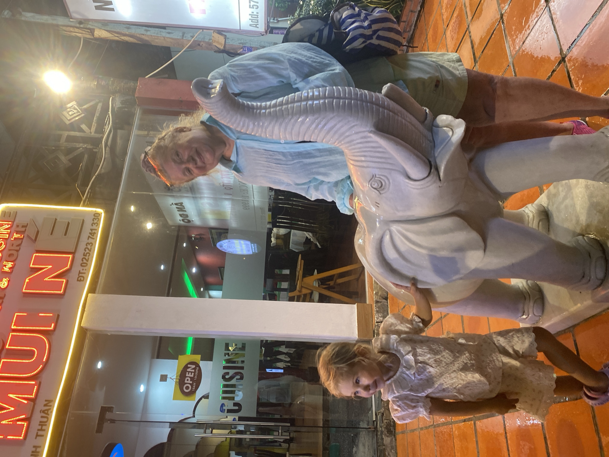 A woman and a young girl stand next to a large white elephant statue in front of a restaurant. The restaurant has a bright neon sign that reads "MUINE NORTH" in red lettering. Below the sign, glass doors reveal the interior of the restaurant, with tables and chairs visible. A yellow "OPEN" sign is hanging on the glass. The ground is tiled with orange squares, and appears wet. The woman is wearing a blue shirt and light-colored shorts, with a striped bag slung over her shoulder. The girl is wearing a white dress with ruffles and lace details, and pink shoes. The elephant statue has its trunk raised. The scene suggests an evening setting, with artificial lighting illuminating the area.