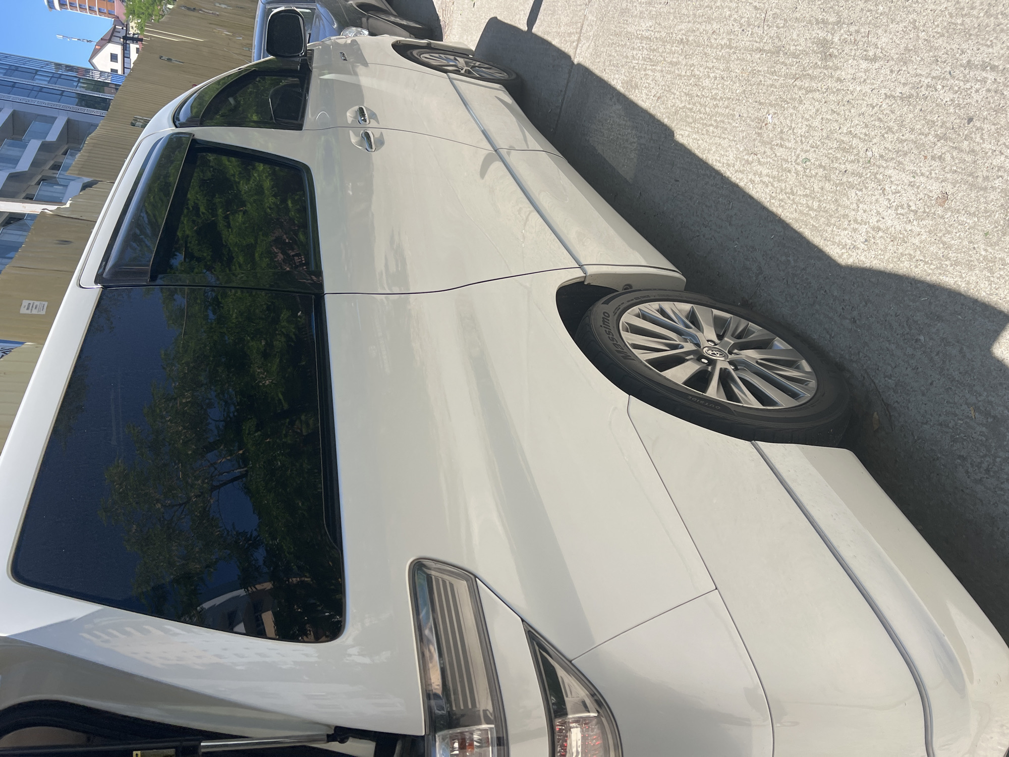 A white Toyota Alphard or similar minivan is prominently featured, parked on a concrete surface under clear, sunny daylight. The vehicle is oriented with its side and rear toward the viewer, showcasing its tinted windows, black window visors, and multi-spoke alloy wheels with a Toyota logo on the hubcaps. The tire sidewall clearly displays the text "OUTSIDE."
Strong, dark shadows are cast across the concrete pavement, suggesting a bright sunny day, likely midday or early afternoon. In the background, beyond a wooden fence, modern multi-story buildings with glass facades are visible, along with a more traditional building featuring a red roof. Reflections of trees and architectural elements are visible in the car's dark windows. A dark-colored vehicle is partially visible parked in front of the white minivan. The scene depicts an urban setting, with the vehicles appearing stationary. No individuals or specific activities are depicted. The overall impression is a parked car in a city environment, consistent with Sochi, Russia.