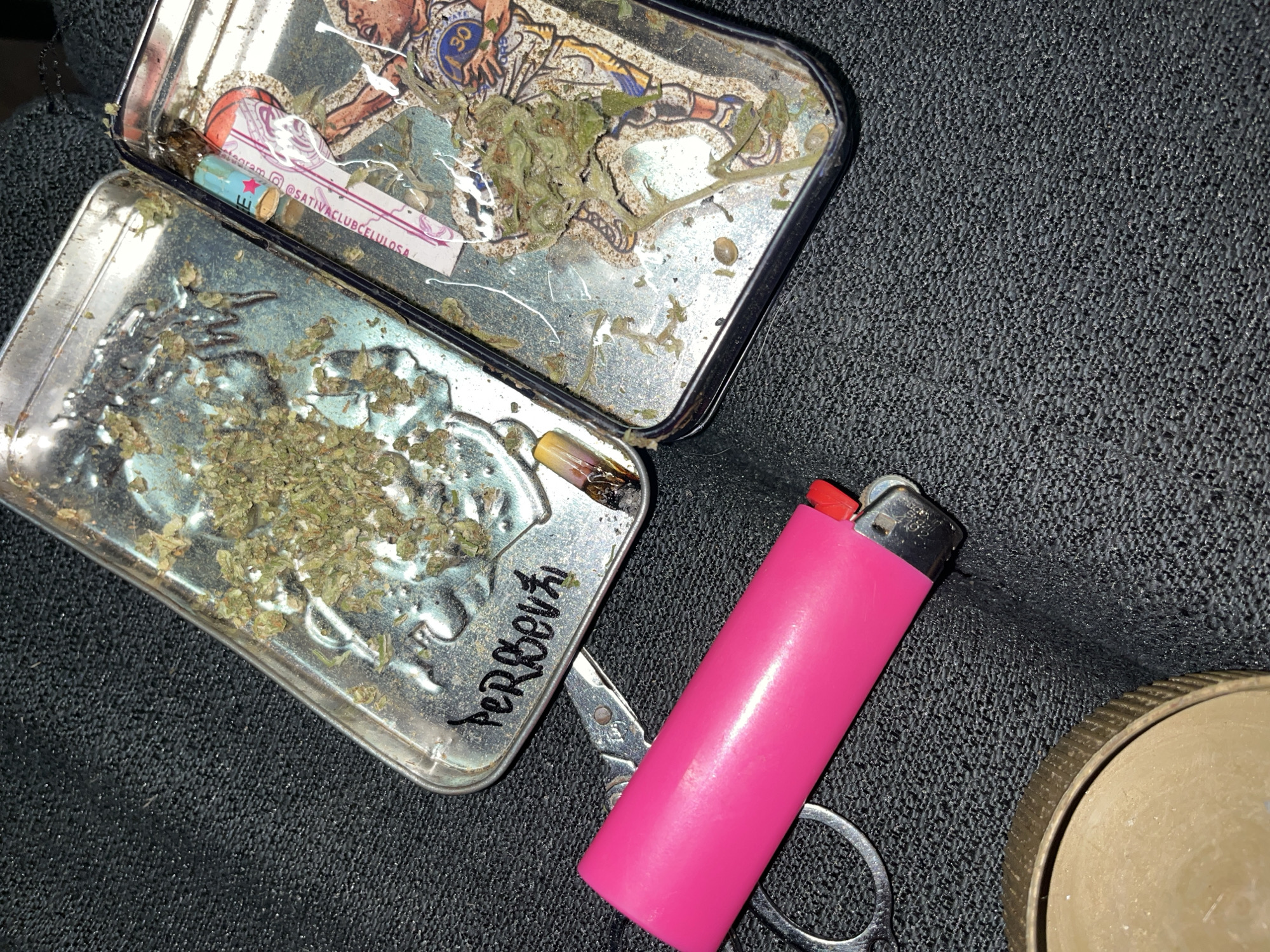 A slightly elevated, angled shot shows an open metal container resting on a dark, textured surface. The container, divided into two sections, holds what appears to be loose cannabis. One section also contains a partially smoked rolled cigarette. Writing is visible on the side of the container. Adjacent to the container is a pink lighter and a pair of scissors. Part of a circular metal object is visible in the lower right corner. The photo was taken in Llavallol, Argentina.