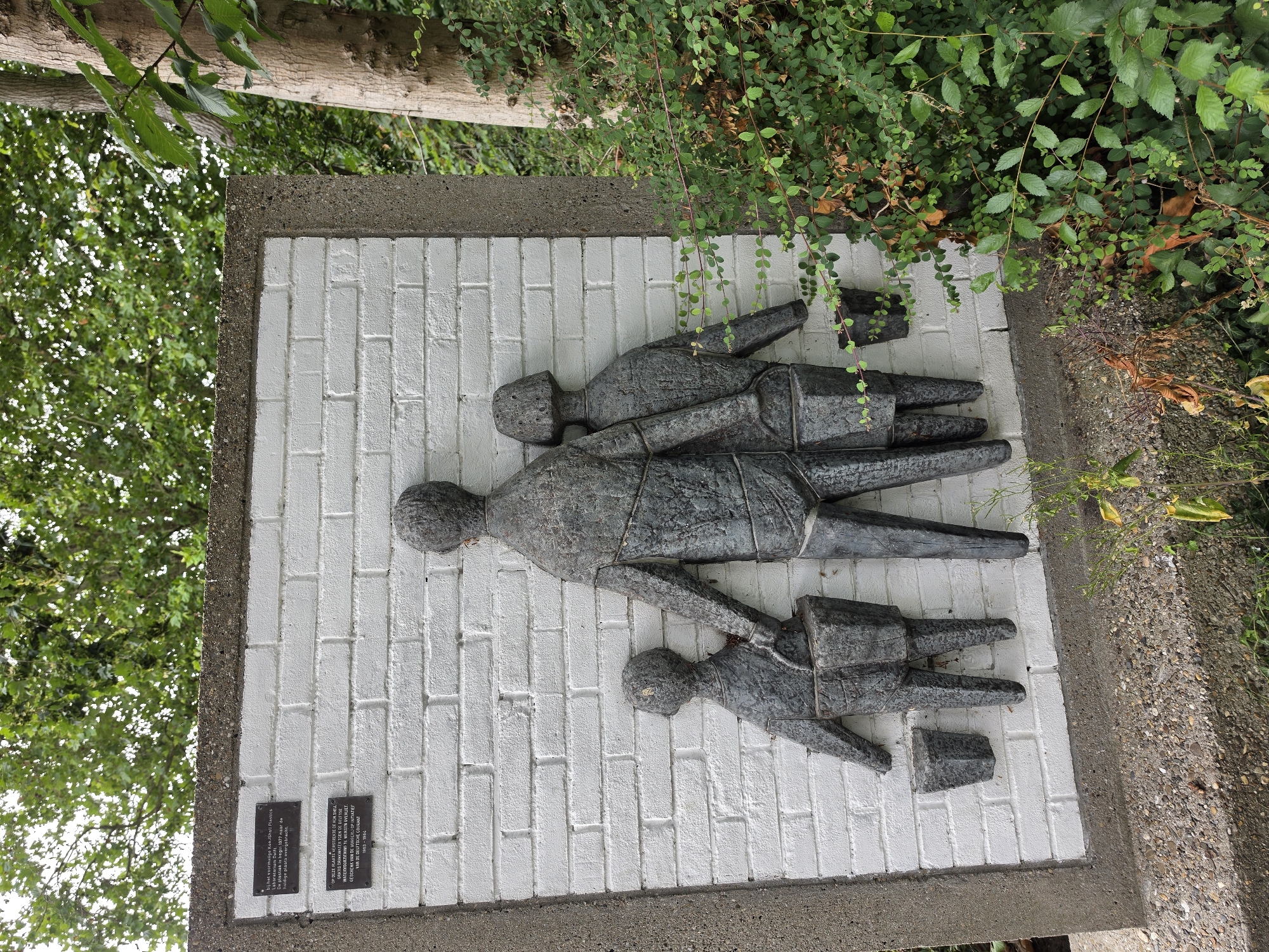This image shows a relief sculpture on a white brick wall, located at the former site of DSM (Dutch State Mines) in Delft, Netherlands. The sculpture depicts three figures: a large central figure, presumably an adult, holding hands with two smaller figures, presumably children. All three figures are carrying buckets. The central figure and the child on the right are holding their buckets at their sides, while the child on the left is holding a bucket that appears to be attached to their waist. The figures are made of a dark, textured material, possibly metal or stone, and are raised from the white brick background.
Two plaques are affixed to the wall above the sculpture. The upper plaque, with a darker background, is partially obscured, but appears to contain text related to the former KanJshi Plastics company and the relocation of a plastic factory in 1977. The lower plaque, also dark, contains text in Dutch, indicating that the relief was created by the Delftse Ondernemers en Ingenieurs (Delft Entrepreneurs and Engineers) and donated by the Delftse Ondernemers Fonds (Delft Entrepreneurs Fund) in 1985.
The sculpture is set within a concrete frame, and the surrounding environment is overgrown with lush green vegetation, including trees and bushes. The lighting suggests it is daytime, with sunlight filtering through the leaves. The overall scene conveys a sense of family or community, with the figures carrying buckets possibly symbolizing work or a communal activity. The presence of the plaques suggests a historical and commemorative purpose for the artwork.