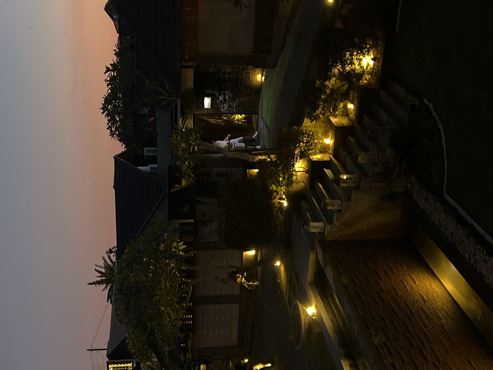 The photograph depicts an outdoor scene in North Kuta, Indonesia at twilight or dusk. A person in light-colored clothing is lying down within a stone archway. The area is illuminated by small, warm-toned lights strategically placed along stairs and in vegetation. There is a building with a dark roof visible on the left. The sky is a gradient of grey to orange. The environment appears to be a garden with trees and shrubs. The scene is rotated 90 degrees.