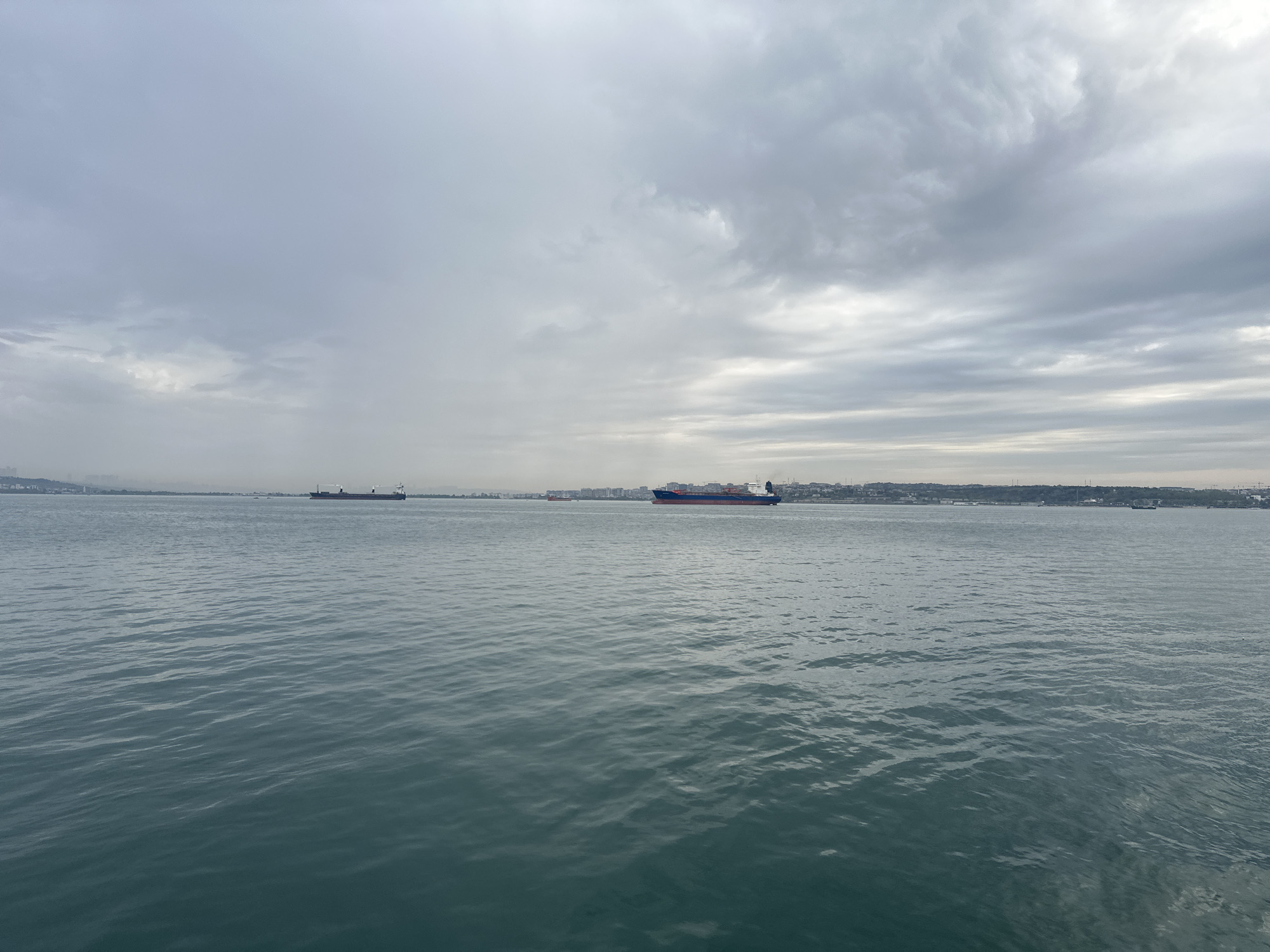 The image depicts a body of water, likely a sea or bay, under a cloudy sky. Several ships are visible in the distance, positioned horizontally across the frame. In the background, a city or urban area extends along the shoreline, obscured by haze and distance. The sky is predominantly gray, with varying cloud formations that dominate the upper portion of the image. The water's surface is rippled, reflecting the ambient light and sky conditions. The image was taken in the city of Unknown, Türkiye.
