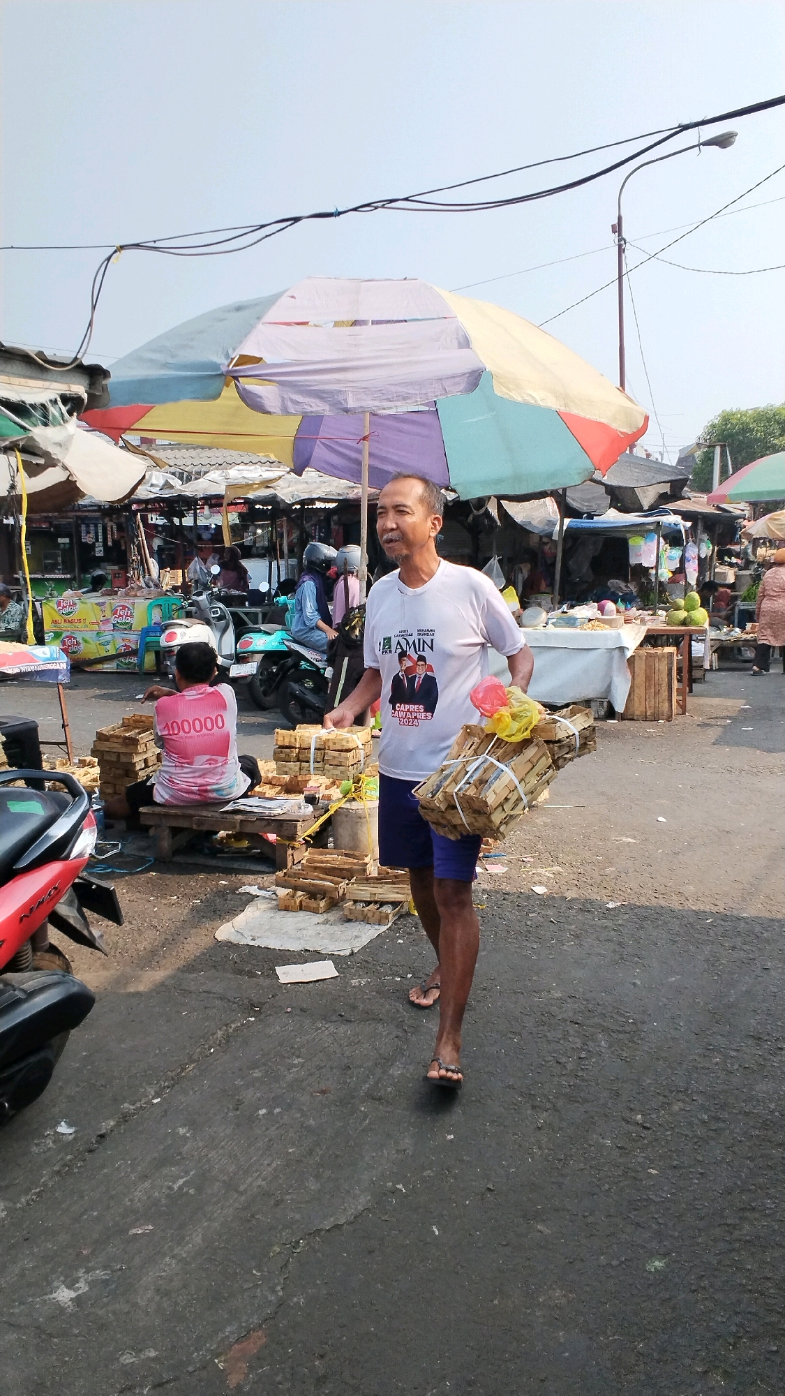 A man walks through an outdoor market in Mojokerto, Indonesia, carrying several bundles of what appear to be packaged goods. He is wearing a white t-shirt with a political campaign graphic featuring images of two men and text that reads "AMIN CAPRES CAWAPRES 2024," blue shorts, and dark flip-flops. In his right hand, he carries a bunch of yellow and red plastic-wrapped items. The market is filled with stalls covered by colorful umbrellas, and there are motorcycles and people in the background. In the foreground to the left, part of a red and black motorcycle is visible. To the left of the walking man, another person sits on a bench wearing a pink shirt with "400000" printed on the back. The ground is paved and appears to be wet in places. The sky is clear and blue, indicating a sunny day.