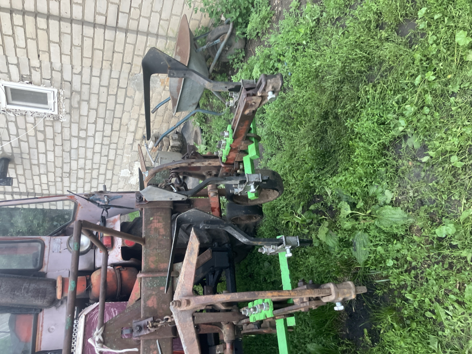 The image depicts agricultural machinery parked outdoors on a grassy area. The machinery appears to be a tractor with attached implements, possibly for plowing or tilling, with several metal components painted in orange and green. A wheelbarrow is visible behind the tractor. In the background, a brick building with a window is present. The overall scene suggests an outdoor setting associated with farming or a rural property. The image was taken in Vovkivka, Ukraine.