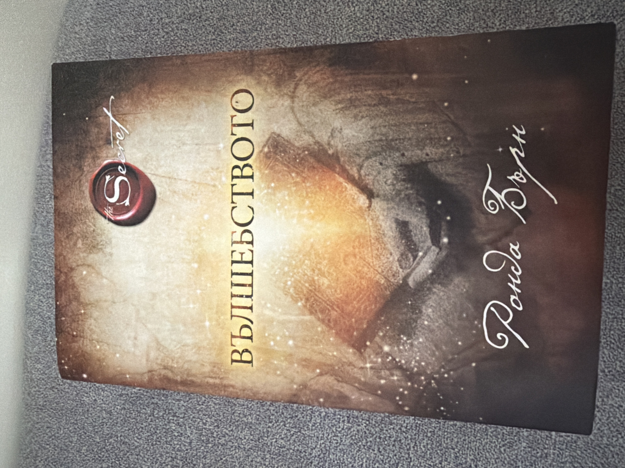 The image shows the cover of the book "The Secret" by Rhonda Byrne, with the title in Bulgarian: "ВЪЛШЕБСТВОТО". The cover features a stylized wax seal with "the Secret" written on it. The background is a textured, sepia-toned image with glowing light and sparkles. The author's name, "Rhonda Byrne" in Bulgarian script, is also visible at the bottom of the cover in white cursive. The book is lying flat on a textured gray surface, likely a carpet or fabric. There is no identifiable city or location within the image itself.
