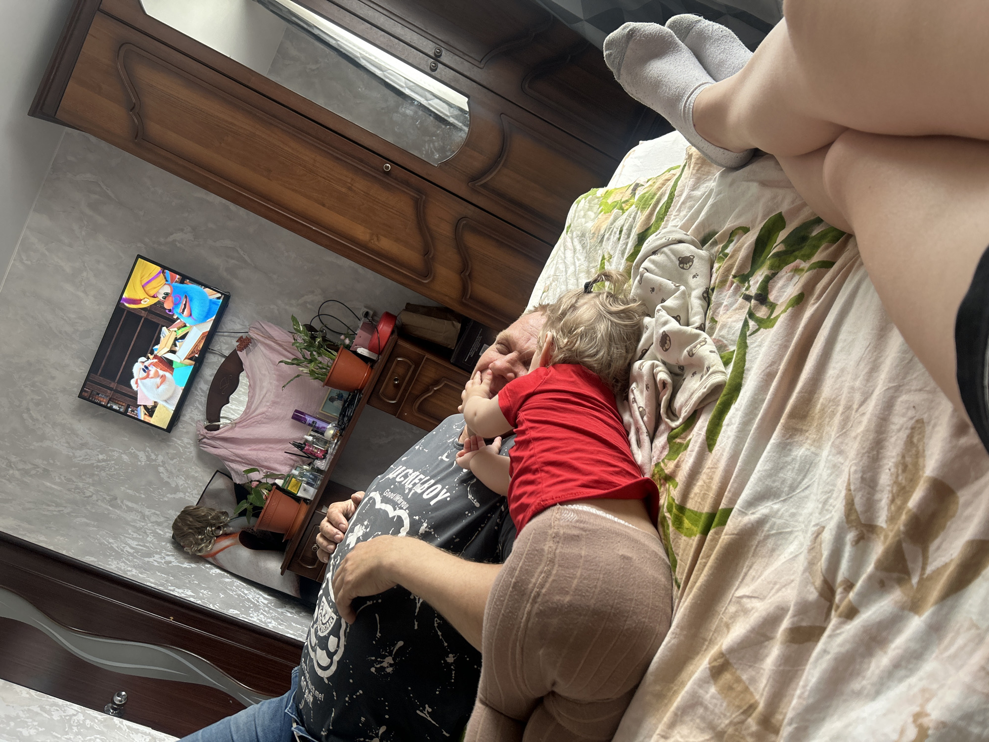 The image shows a man and a young child on a bed in a room. The child is wearing a red shirt and light brown leggings and appears to be interacting with the man. The man is wearing a dark t-shirt with a white design and jeans. A person's legs in white socks are also visible on the bed. In the background, there is a television on the wall displaying a cartoon, a dresser, and a decorative shelf with plants and other items. The room has a light-colored wall with a patterned design. There is no indication of location.