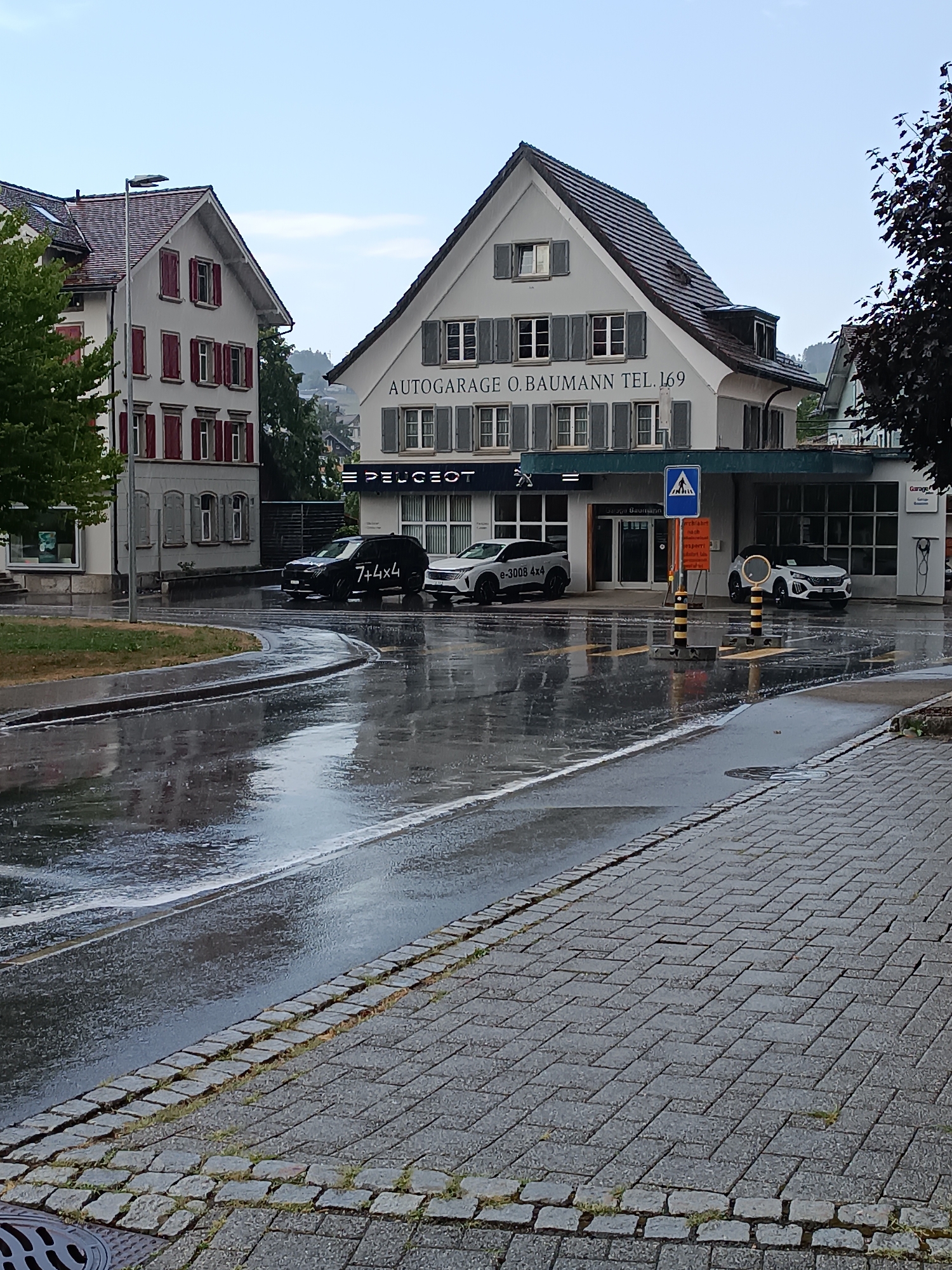 The image captures a street scene in the city of Appenzell, Switzerland, on a rainy day. In the foreground, a wet asphalt road curves to the left, reflecting the overcast sky and surrounding buildings. Cobblestone pavement lines the right side of the road, with patches of grass growing between the stones.
Dominating the midground is a car dealership, identified by the signage "AUTORARAGE O. BAUMANN TEL. 169" and "PEUGEOT". Two cars are parked in front of the dealership: a black SUV with "7+4x4" written on its side, and a white SUV, possibly a Peugeot e-2008 4X4, with "e-3008 4X4" visible on its door. A blue pedestrian crossing sign with a white figure stands near the entrance. Traffic barriers, black and yellow striped, are positioned on the road.
To the left of the dealership, a traditional Swiss building with a light-colored facade, red shutters, and a dark tiled roof is visible. Another building, partially obscured by trees and the dealership, is seen further down the street. The sky is overcast, suggesting continued rainfall. The overall atmosphere is damp and quiet, characteristic of a rainy day in a European town.