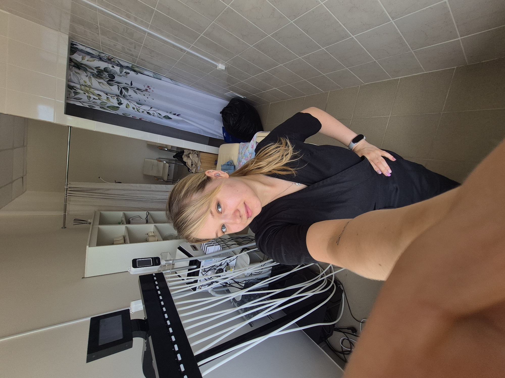 A young woman with blonde hair is taking a selfie in a room that appears to be a medical or spa office, possibly located in the city of Chaikovsky, Russia. She is wearing a black V-neck scrub top and a silver smartwatch. In the background, there is medical equipment, including a large unit with multiple white tubes, a monitor, and shelves filled with various items. A patterned shower curtain is visible on the right side of the image, and a large black trash bag is on the floor. The walls are tiled in a neutral tone, and the floor is covered with beige tiles. The overall impression is of a clean, functional, and somewhat clinical environment.