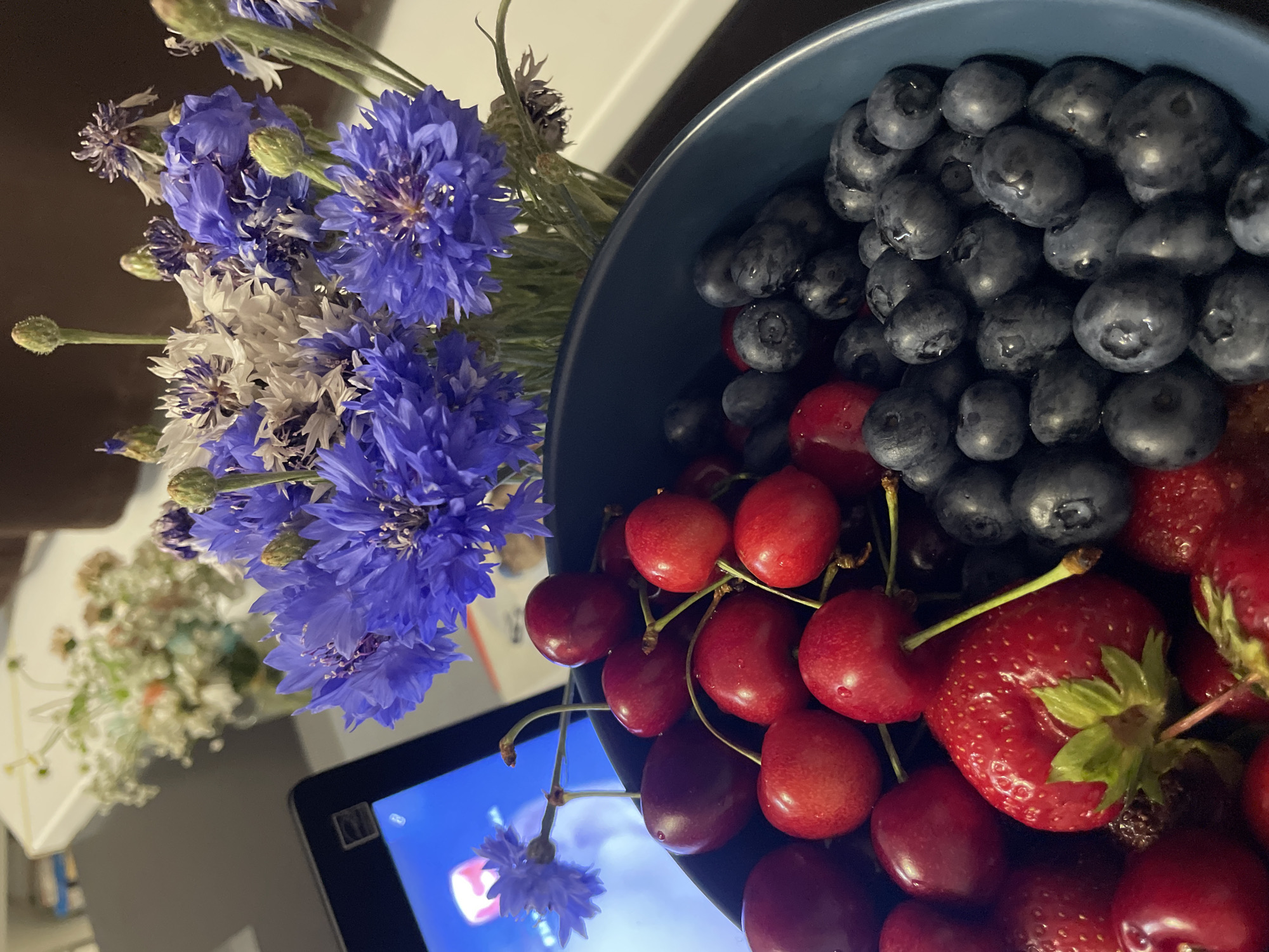 The image shows a close-up view of a blue bowl filled with fresh fruit, including cherries, blueberries, and strawberries. In the background, a bouquet of blue and white cornflowers is visible, along with a tablet screen displaying a colorful image. The overall impression is one of a vibrant and refreshing arrangement, possibly set on a table. There is no indication of the city of Sochi, Russia, or any specific location within it. The scene is static and focuses on the still life composition.