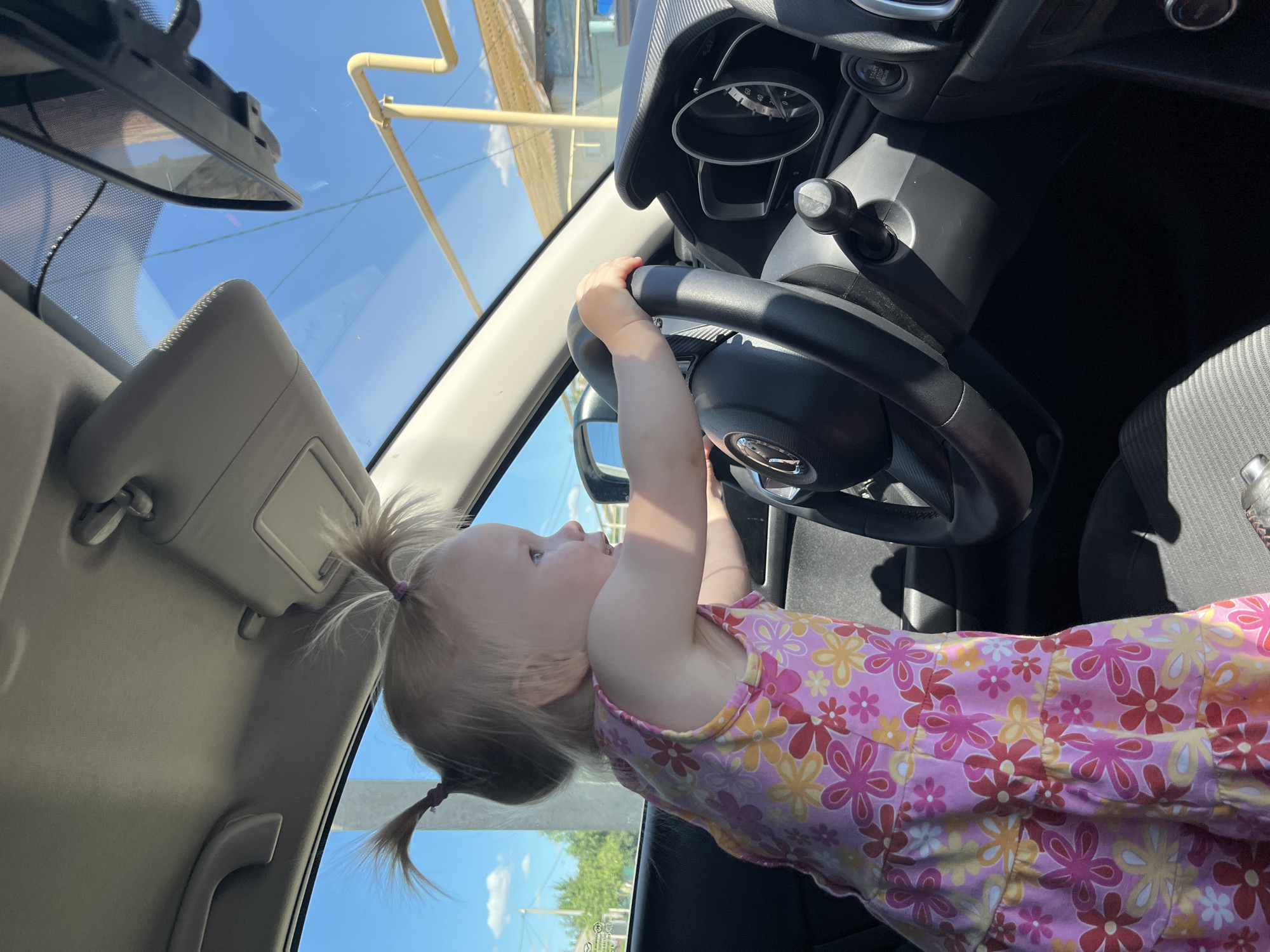 A young blonde child with two pigtails, wearing a pink floral dress, is sitting in the driver's seat of a car, holding onto the steering wheel. The child's face is turned upwards and to the left, with a happy expression. The scene is captured from a low angle, looking into the car. The interior of the car is visible, including the dashboard, steering wheel, and part of the passenger seat. Outside the car, the sky is bright blue with some white clouds, suggesting it is daytime. There are some blurry structures visible through the windshield, possibly buildings or scaffolding. A sun visor is visible in the top left corner of the image. The sunlight is strong, casting shadows within the car. The child's hands are gripping the steering wheel as if they are driving. The overall impression is one of playful curiosity, with the child exploring the car. The provided location context is Dubovka, Russia.