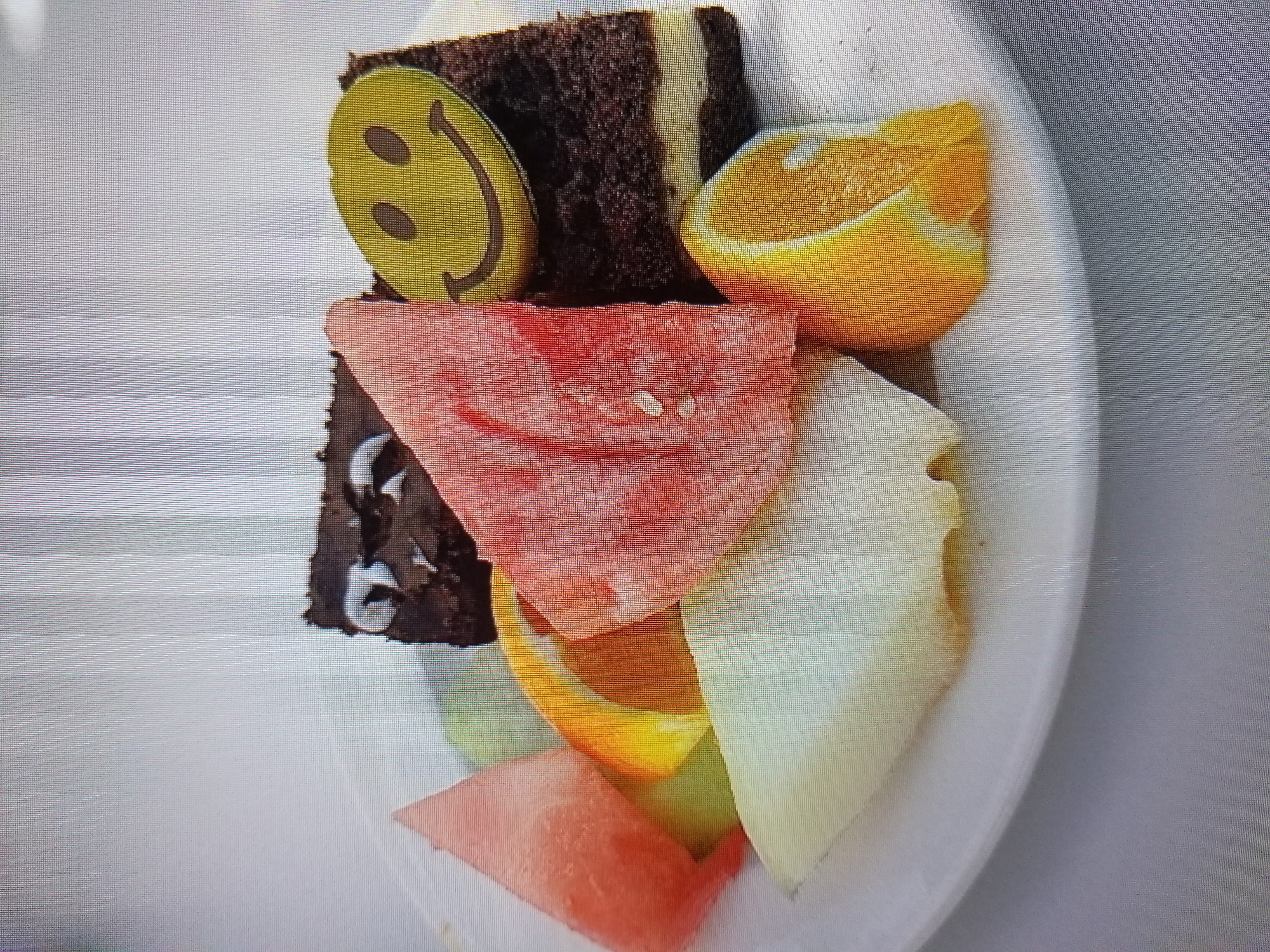 A white plate holds a slice of dark chocolate cake with a yellow smiley face decoration, several slices of watermelon, a slice of orange, and a piece of light-colored fruit, possibly pear or melon. The food is arranged on a white surface. The image appears to be taken from a slightly overhead angle. The scene does not contain any identifiable landmarks or elements to associate it with a specific city or location like Tiraspol, Moldova.