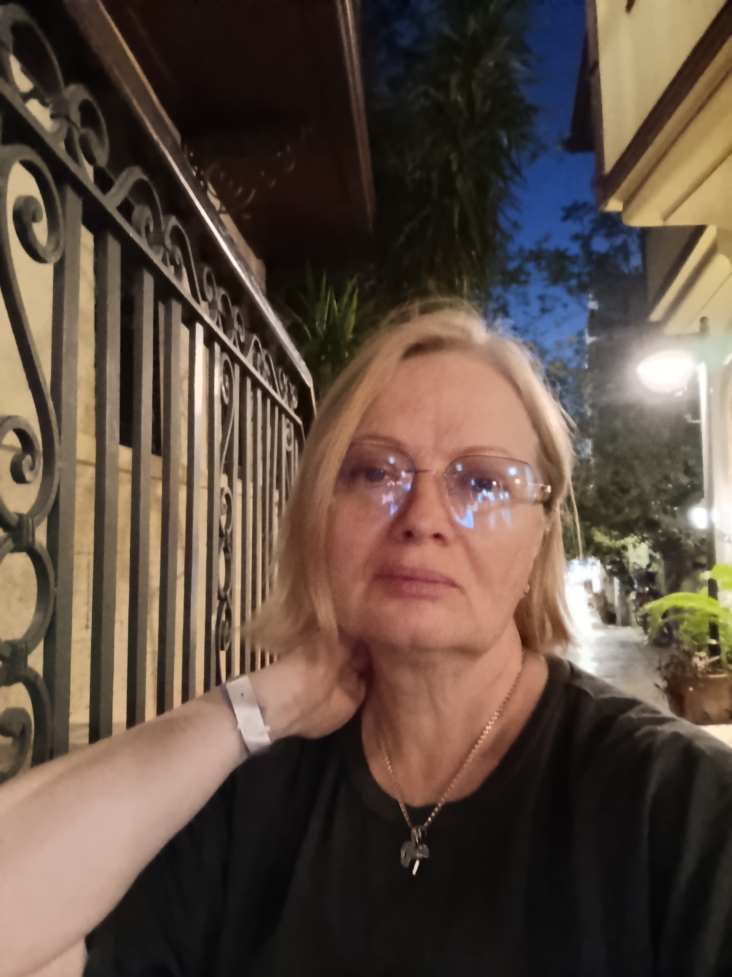 A middle-aged Caucasian woman with blonde hair and glasses is taking a selfie. She is wearing a dark t-shirt and a necklace. Her left hand is raised to her neck and she is wearing a white wristband. The background shows an outdoor evening scene. To the left, there is a black metal fence with decorative scrollwork. Behind the fence, there are vertical bars. In the distance, a street is visible with plants and lights. The sky is dusky blue. The location is likely in Antalya, Turkey, based on visual characteristics.