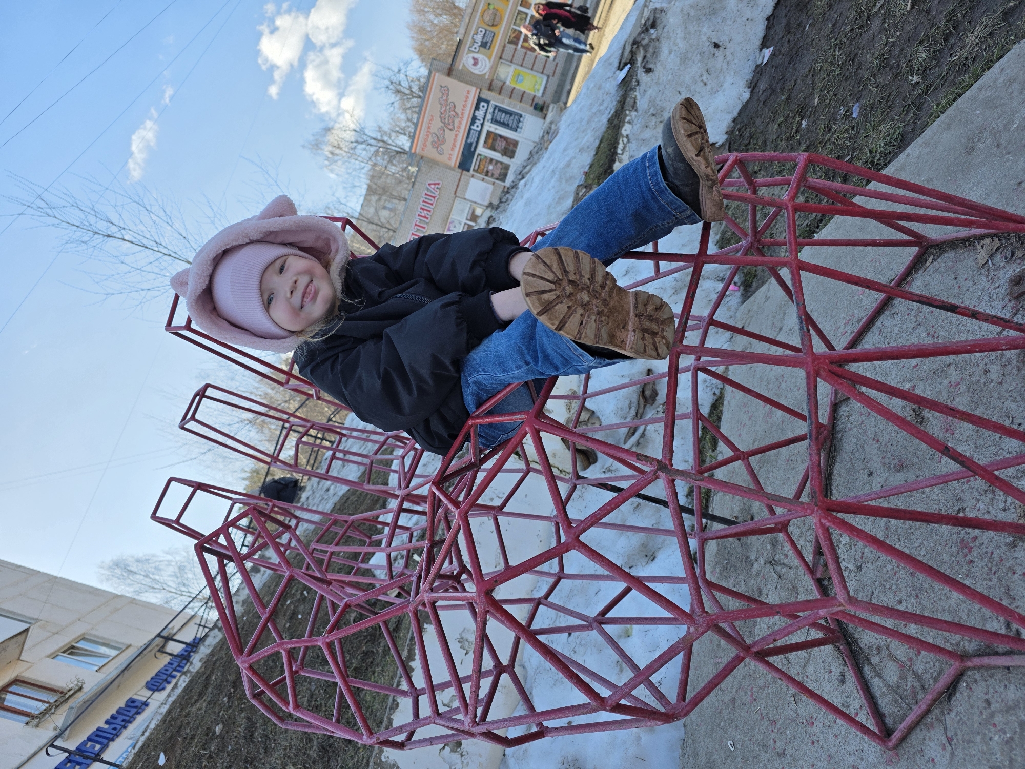 A young girl sits atop a red metal sculpture, likely in a public space in the city of Chaikovsky, Russia. She wears a pink hat with ears, a black jacket, and blue jeans, her brown boots extending outward. The sculpture's geometric design suggests a playful structure. In the background are buildings, bare trees, patches of melting snow, and passersby walking near stores, providing a context of early spring in a populated area.