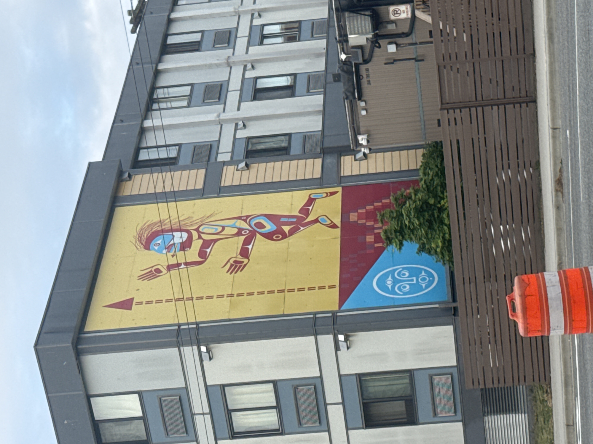 A daytime eye-level view shows the side of a tall building with a large mural depicting an Indigenous design, located in Vancouver, Canada. The building is primarily gray and white, with multiple windows. The mural, in shades of yellow, red, and blue, dominates the center of the frame. It shows a stylized figure with an arrow pointing towards it. The right side of the mural is decorated with a distinct blue design. A green bush partially covers the right edge of the mural. A brown slatted fence runs along the right side of the building and parallel to the street, and an orange-and-white traffic barrel is positioned on the street. Overhead wires are visible against the sky.