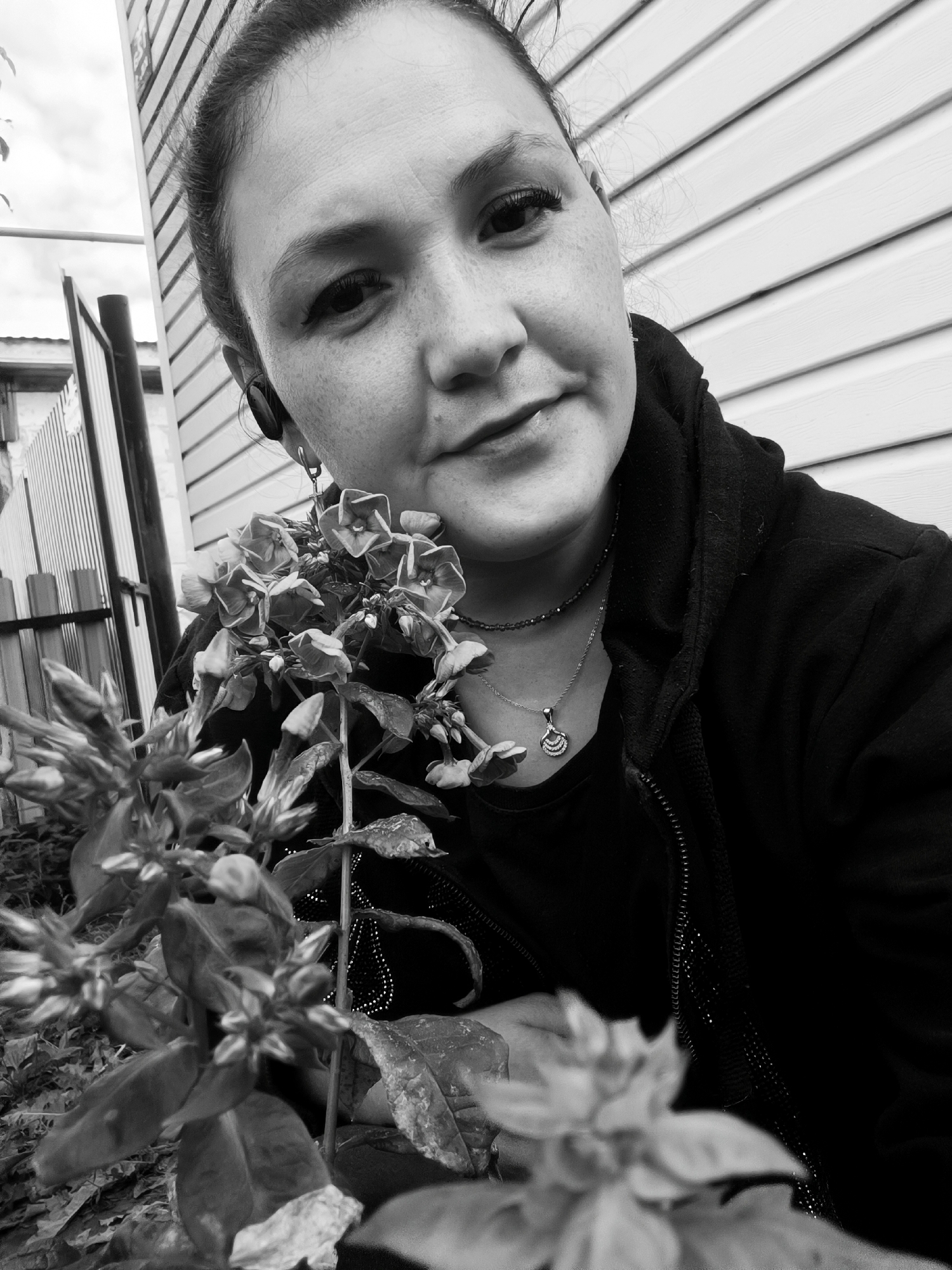 A black and white, medium shot, selfie-style photograph shows a woman in an outdoor setting. She is positioned in the lower right quadrant of the frame, facing slightly to her left. The woman has her hair pulled back in a bun. She is wearing a black zip-up hoodie over a black t-shirt and a necklace with a shell pendant. She also has an earbud in her left ear and a hoop earring. In the foreground, to the left, are several blooming flowers and leaves. In the background, to the left, is a metal fence and part of a building with white horizontal siding. The sky is visible in the upper left corner. The overall scene suggests a casual, outdoor setting. The provided hints indicate the location is in the city of Karsha, Russia.