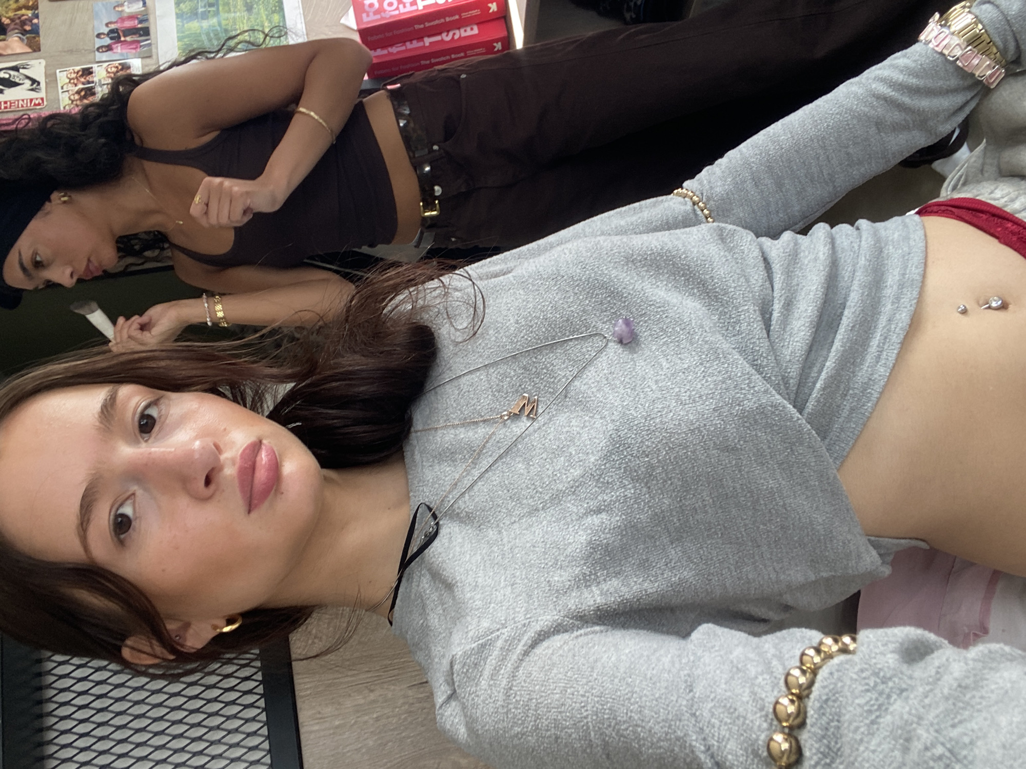 Two women are visible in an indoor setting. One is wearing a brown tank top and matching brown pants with a belt, a black beanie, and jewelry, including bracelets. The second woman is wearing a gray long-sleeved shirt, a necklace, bracelets, and has a visible belly button piercing. Books are visible on the surface where the women are sitting. The location in the city of London, United Kingdom is not identifiable.
