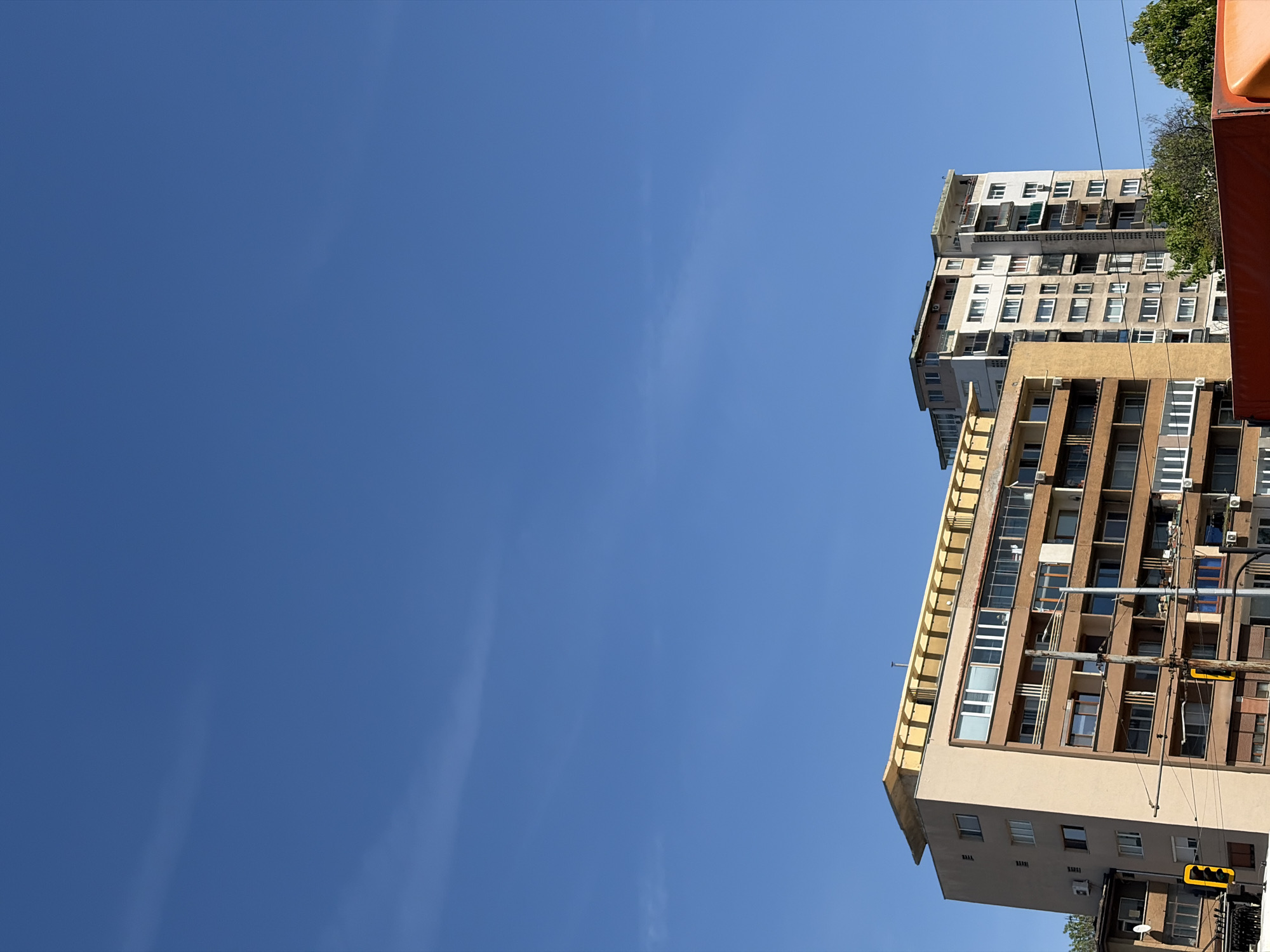 The image, rotated 90 degrees counter-clockwise, captures a daytime urban scene under a vast, clear blue sky. The sky, occupying the majority of the left side of the frame, is a deep blue with only faint, wispy clouds visible, suggesting bright and sunny weather.
On the right side, multiple high-rise buildings dominate the frame. The closest building in the foreground is multi-storied, featuring a facade primarily composed of light brown and beige panels, interspersed with darker brown sections. It has numerous windows, many with external shutters, and balconies. Architectural details with a yellowish hue are visible along its top-left edge. Several air conditioning units are mounted on its exterior. Behind this structure, another tall building with a lighter, off-white or grey facade can be seen, also characterized by multiple windows.
Green foliage from trees is visible at the very top and bottom right edges of the frame, indicating the presence of greenery in the urban setting. Power lines are visible stretching across the upper right portion of the sky. A yellow traffic light, oriented horizontally, is discernible near the bottom right section of the foreground building.
The scene depicts a typical urban environment, likely a street view looking upwards at residential or office blocks. There are no people visible, and no discernible text is present on any of the structures or signs. The architectural style is consistent with that found in Eastern European cities, aligning with the context of Sofia, Bulgaria.
