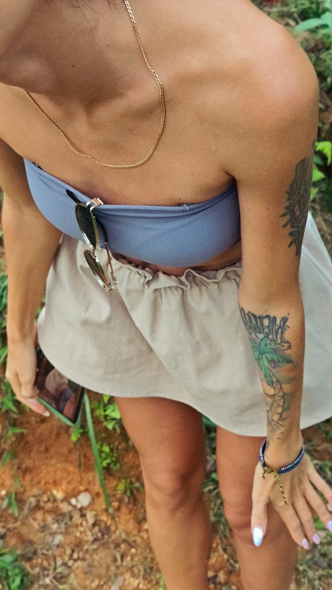 An overhead, close-up shot captures a person, likely female, from the chest to the upper thighs, situated outdoors in what appears to be Nậm Dịch Commune, Vietnam. The person has tanned skin and is dressed in a light blue ribbed strapless top and a light beige ruffled skirt or shorts. A thin gold chain necklace adorns their chest, and a pair of sunglasses is hooked onto the top.
Both arms display tattoos. The left forearm features a vibrant design including a green palm tree, above which text resembling "NÀNG BIỂN" is visible. The upper right arm has a darker, intricate pattern. A dark blue beaded bracelet with a green/gold string is worn on the right wrist, and the fingernails are painted with an iridescent light purple/blue polish. The person is holding a smartphone in their left hand, its screen reflecting light.
The setting is an outdoor, natural environment, characterized by reddish-brown earth and vibrant green foliage and grass in the background. The bright, clear lighting suggests it is daytime with warm, sunny weather. The posture indicates the person is either standing or leaning forward, perhaps looking down at the ground or their phone.