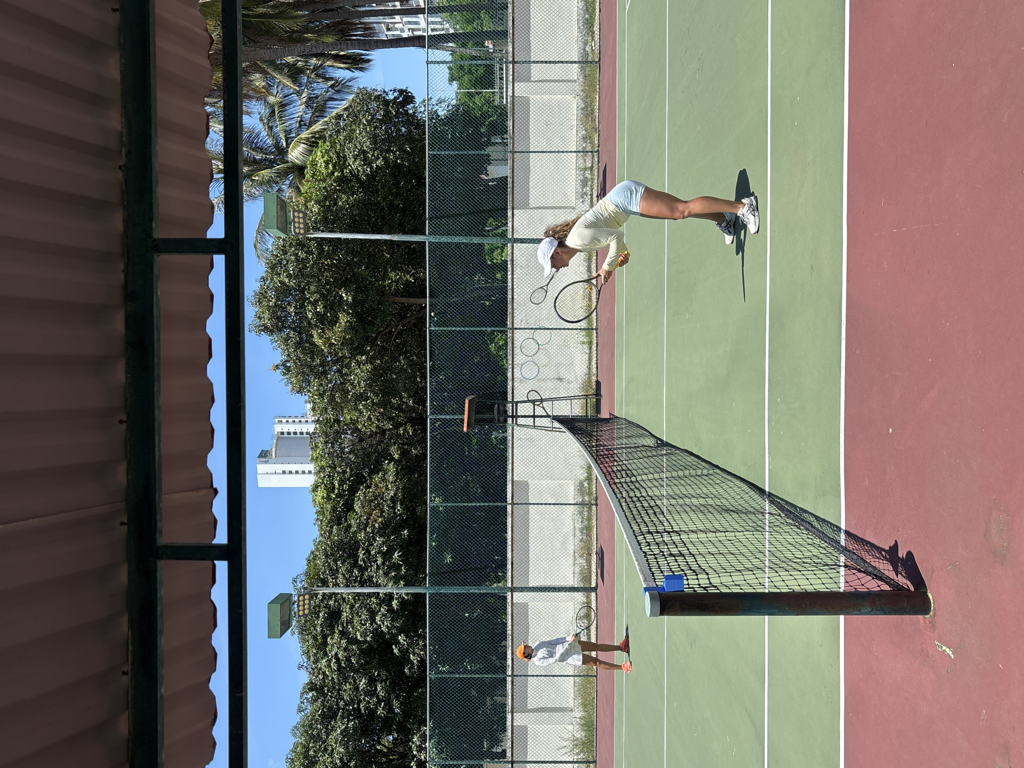 This image shows a tennis court in Nha Trang, Vietnam, likely at a resort or club. Two young women are on the court, dressed in tennis attire and holding rackets. The woman on the left is wearing a white long-sleeved shirt, white shorts, and an orange visor. She is positioned near the baseline of the court, holding her racket in a ready stance. The woman on the right, closer to the net, is wearing a light yellow long-sleeved shirt and light blue shorts. She is in a lower stance, also holding her racket as if preparing to receive a serve or play a shot. The court surface is green with white lines, and the surrounding area includes a net, a umpire's chair, a chain-link fence, lush green trees, and palm trees. In the background, tall buildings are visible, suggesting an urban or semi-urban setting. The sky is clear and blue. The image appears to be taken from under a covered area, possibly a spectator shelter or part of a building, indicated by the corrugated metal roof structure at the top of the frame. The lighting suggests it is daytime, with bright sunlight casting shadows on the court.