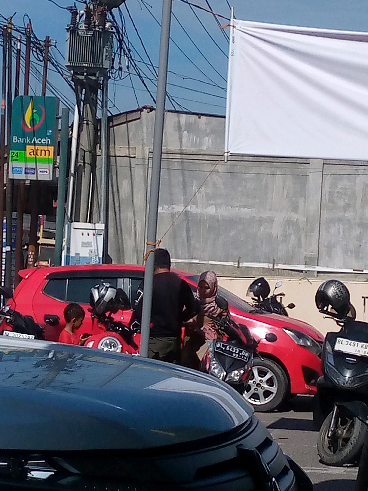This is a street-level, medium-angle shot captured in Blangpeuria, Indonesia, on a bright, sunny day. In the foreground, the hood of a dark-colored vehicle reflects the sky and surrounding elements. The midground features a bright red hatchback car parked on the street, partially obscured by a parked black motorcycle in the right foreground. A young boy in a red shirt is visible standing next to the red car. To the left of the red car, another black motorcycle is parked, and a woman wearing a hijab is seated on it. Behind the red car, a man in a black shirt and green shorts is standing. The background reveals a concrete wall, a utility pole with transformers and numerous electrical wires, and a large white banner suspended from above. A sign for "Bank Aceh" with an ATM symbol is also visible on the utility pole. The scene appears to be an outdoor public space, possibly near a bank or commercial area, with vehicles and pedestrians present.