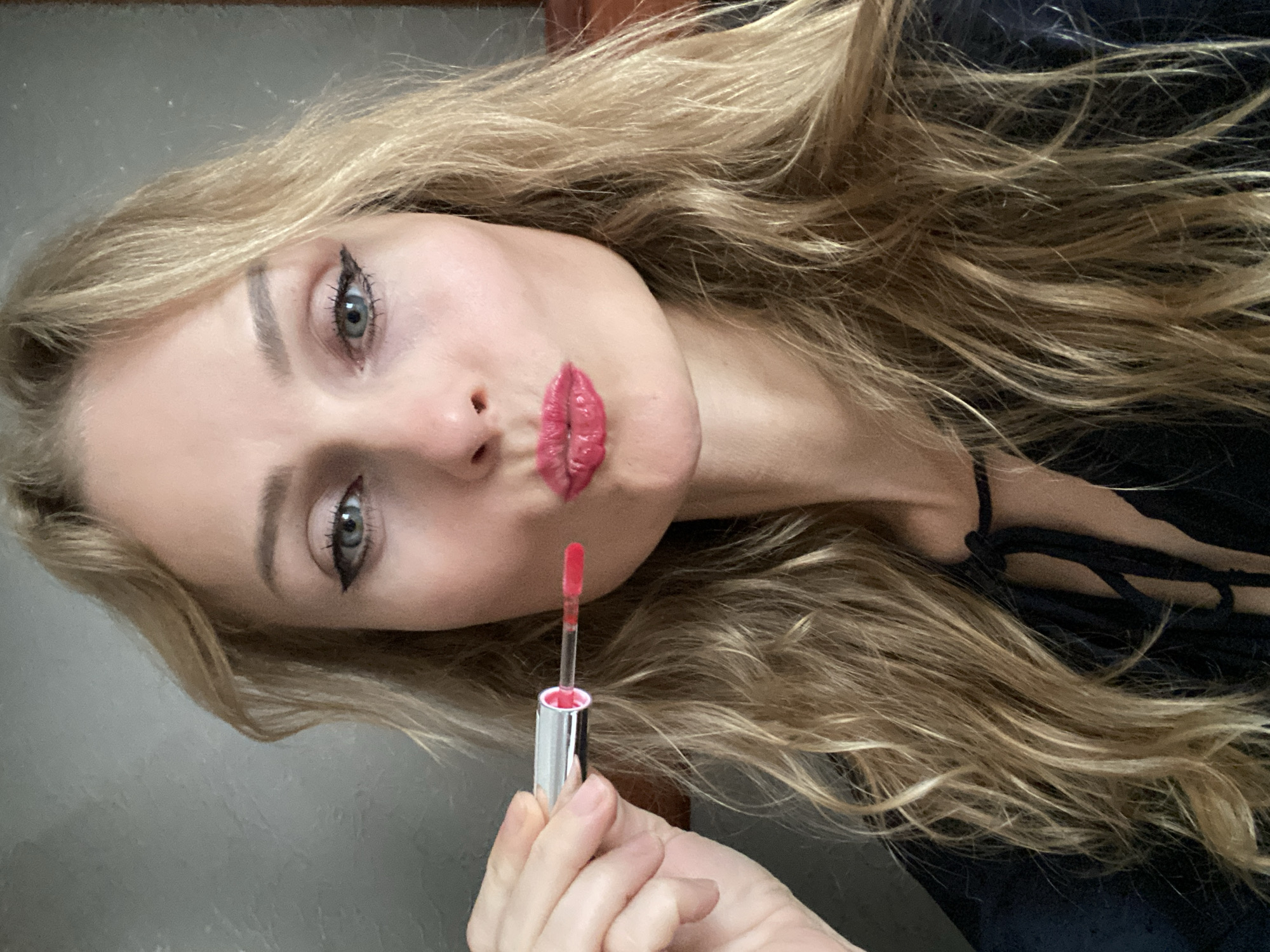 A woman with blonde, wavy hair is applying bright pink lipstick from a silver tube. Her lips are pursed, and she is looking into the camera. Her eyes are green with black eyeliner. She is wearing a dark top with a tie detail at the neck. The background is a plain, textured gray wall. There is no discernible indication of the city of Lida, Belarus.