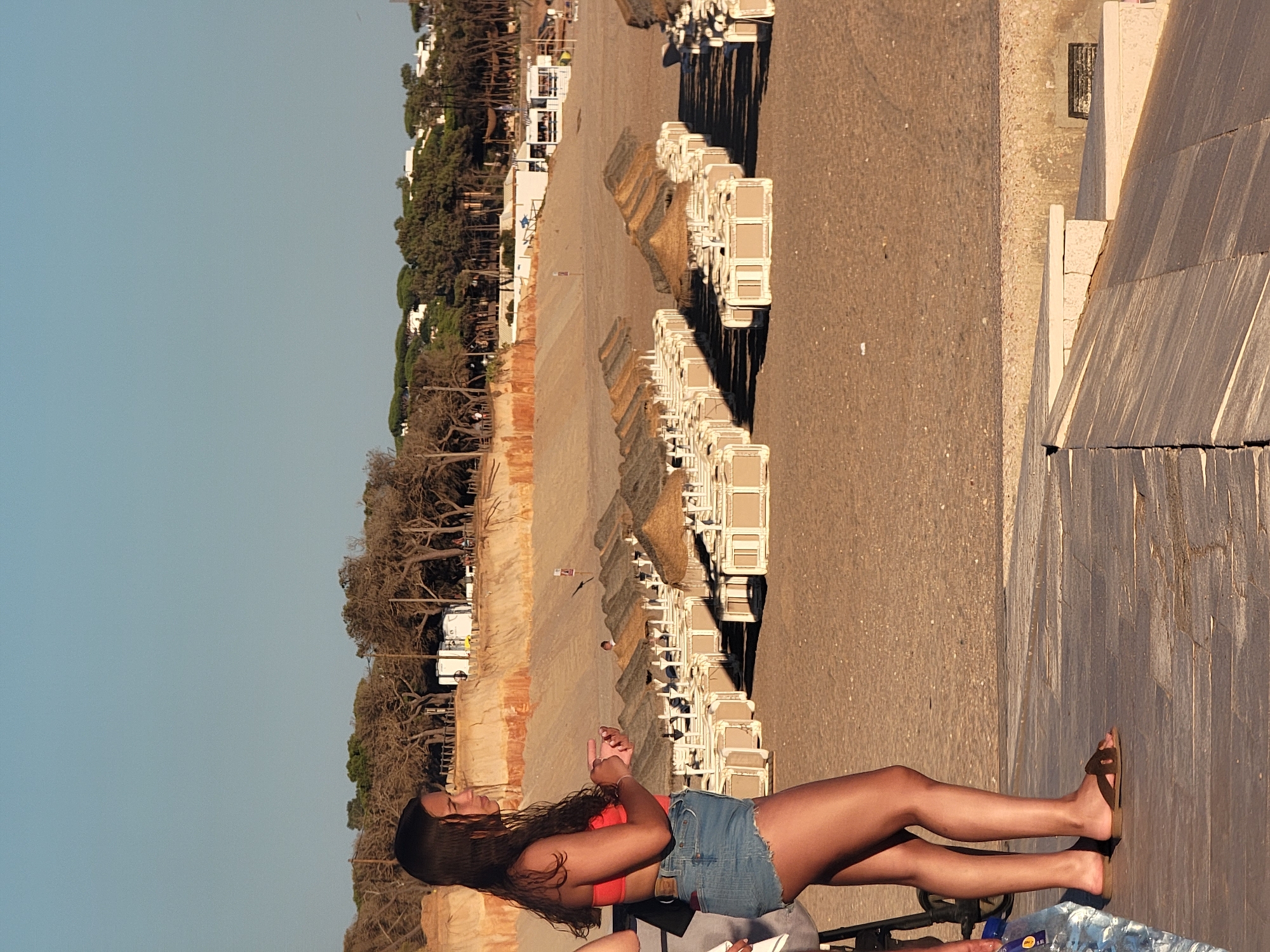 A young woman with long, dark hair stands on a wooden boardwalk at the beach in Quarteira, Portugal. She is wearing an orange bikini top and denim shorts, and is looking to her right. Behind her, rows of empty beach chairs and straw umbrellas stretch across the sand towards a line of trees and buildings on a bluff. The sky is clear and blue. In the foreground, to the left of the woman, part of a stroller and a plastic bottle of water are visible. The lighting suggests it is late afternoon.