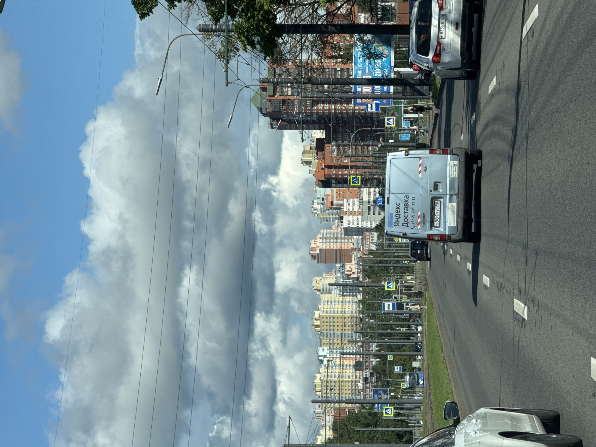 The image depicts a street view in Saint Petersburg, Russia, on a partly cloudy day. The foreground features a road with lane markings and several vehicles, including a white car on the left, a light blue delivery van in the center with "Яндекс Доставка" (Yandex Delivery) written on its side, and a silver SUV on the right. A white van, also with "Яндекс Доставка" visible, is in the center of the frame. In the middle ground, there are pedestrian crossings, traffic lights, and residential buildings with colorful facades, characterized by a mix of yellow, pink, and orange hues. The background shows a skyline dominated by apartment complexes, some with distinctive architectural features. Overhead, power lines stretch across the scene, and streetlights are visible along the road. A few pedestrians are present on the sidewalk. The overall environment suggests a residential or mixed-use urban area.