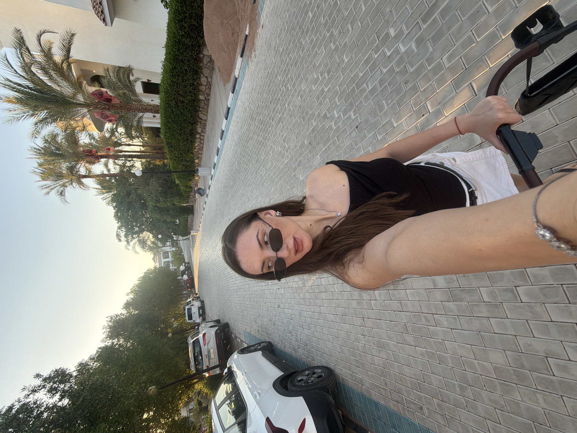 A woman with long brown hair, wearing sunglasses, a black off-the-shoulder top, and white shorts, is holding the handle of a stroller and looking to her left. She is on a paved road lined with parked cars and palm trees. The scene appears to be in a resort area in Sharm El Sheikh, Egypt, with white buildings visible in the background. The lighting suggests it is late afternoon or early evening.