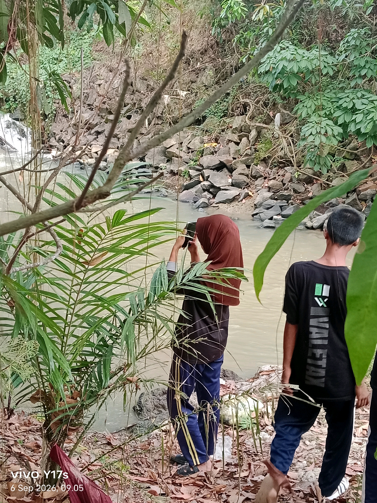 Two individuals are positioned near a body of water, which appears to be a river or stream with a small waterfall visible on the left. The area is dense with foliage, including trees, vines, and leafy plants. Rocks and debris are scattered along the bank. One person, wearing a brown hijab and dark clothing, is holding a device, possibly a camera or phone, up to their face, as if taking a picture. The second person, facing away from the camera, wears a black t-shirt with a white and green graphic and dark pants. The lighting suggests it is daytime, with natural light filtering through the dense vegetation. The scene is set in a natural, outdoor environment, likely a rural or semi-natural area within the vicinity of Rama Kelandungan, Indonesia, given the provided metadata.