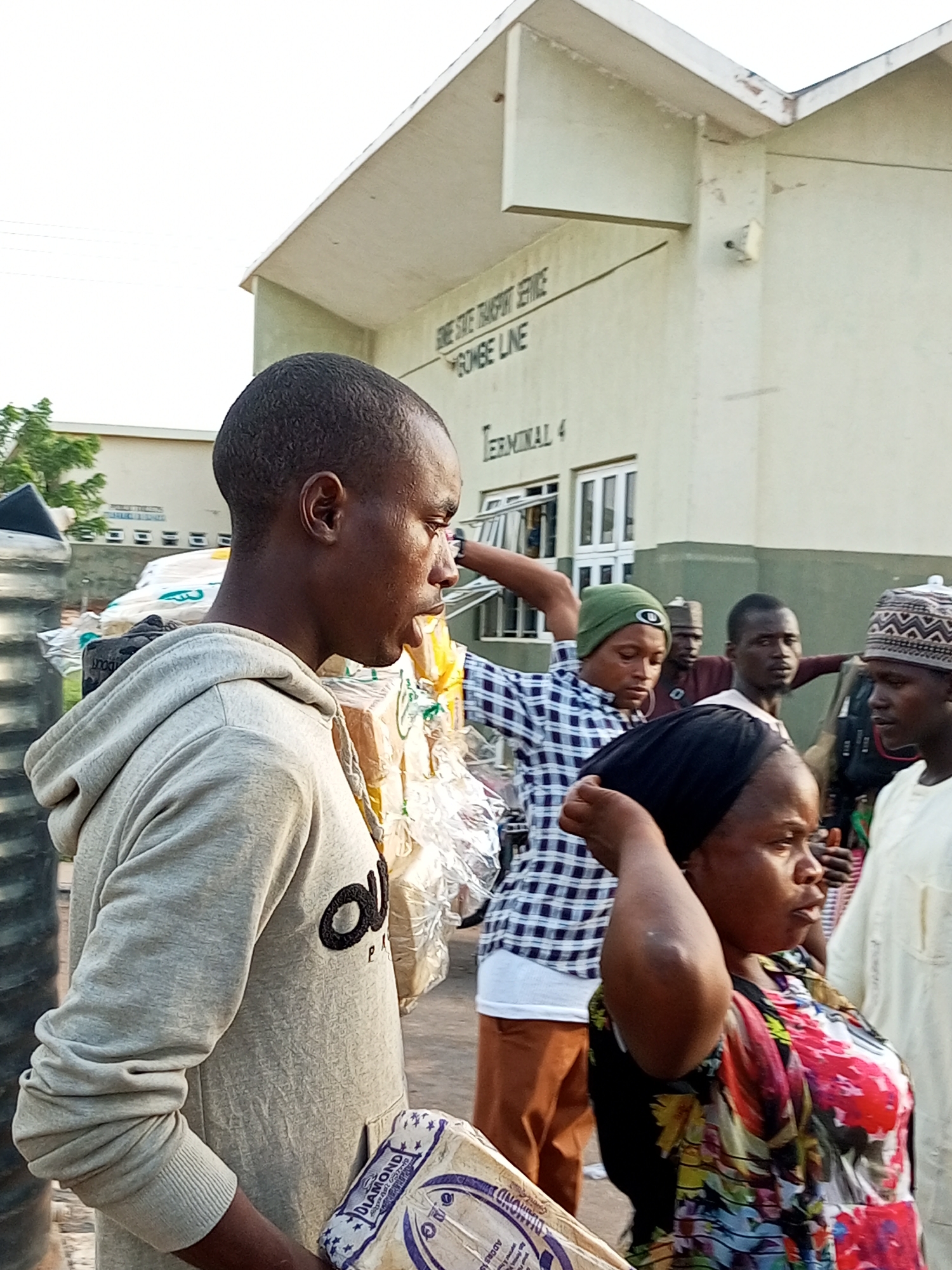 The image depicts an outdoor scene at Terminal 4 of the Gombe State Transport Service, Gombe Line, in Gombe, Nigeria. A group of people are gathered around what appears to be a transportation hub, with several individuals holding or standing near packages. In the foreground, a man wearing a grey hoodie is shown in profile, looking to his right, with a package labeled "DIAMOND" in his hands. Behind him, a woman in a brightly colored floral dress adjusts her headscarf. Other individuals, some wearing traditional attire and headwear, are visible in the background, suggesting a bustling environment. The building in the background features signage indicating the location. The overall atmosphere suggests a moment of departure or arrival, with people preparing to travel or having just arrived.