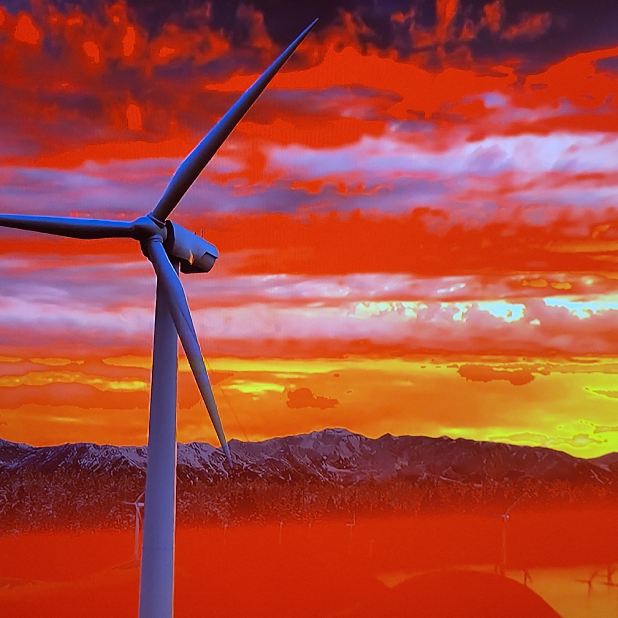 A wind turbine stands prominently in the foreground against a dramatically colored sunset sky. The sky transitions from deep red at the top to yellow and orange near the horizon. In the background, a mountain range is visible. Several other wind turbines, smaller and farther away, dot the landscape, suggesting a wind farm. This image is NOT located in Yerevan, Armenia.