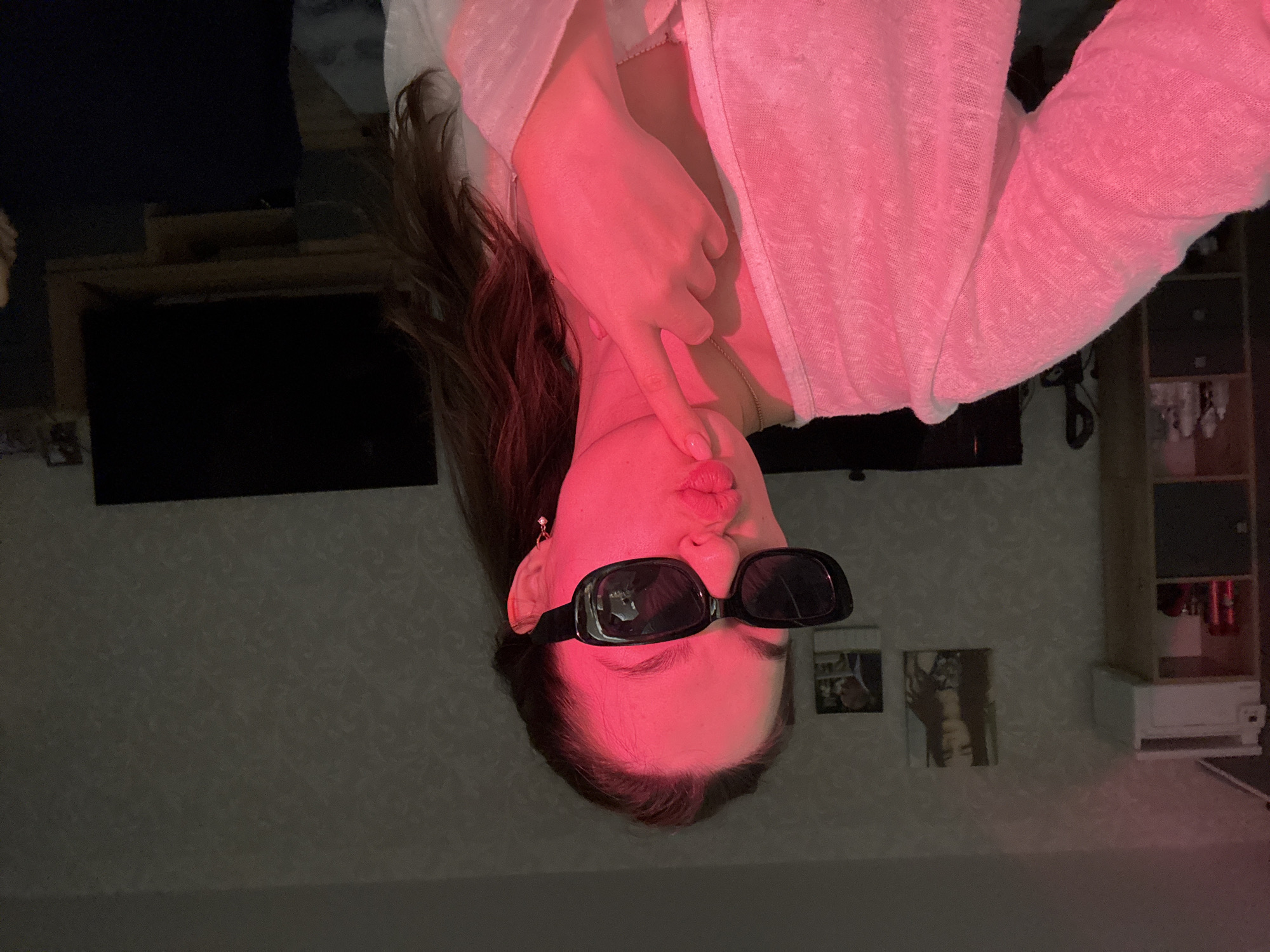 A young woman, illuminated by red light, poses for a selfie in a room. She wears black, rectangular sunglasses and a light pink cardigan over a white garment. Her right hand is raised to her chin, and her lips are pursed. In the background, a shelf unit with various items, including red cans and decorative objects, is visible on the left. A television screen is present on the right. The wall behind her has a textured, patterned wallpaper. The scene appears to be an indoor residential setting, potentially a living room or bedroom. No specific location in Chaikovsky, Russia, is identifiable from the image.