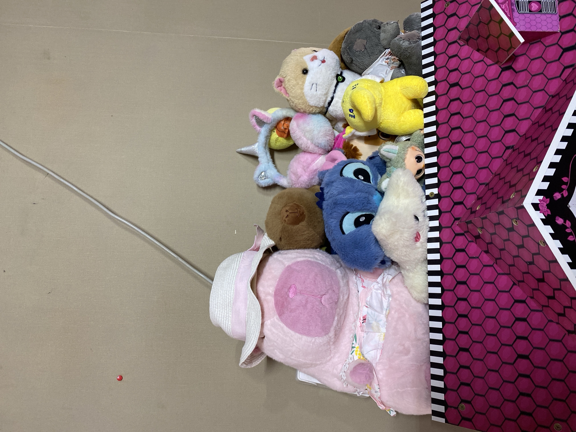 The image displays a collection of stuffed animals piled inside a pink and black hexagonal patterned box. A large pink bear wearing a straw hat with a pink ribbon is prominent in the front left. To its right are several other stuffed animals including a blue Stitch character, a yellow duck-like toy, a tan and white cat, and a gray bear. Various smaller stuffed animals and accessories such as a unicorn headband are also visible. The background is a plain beige wall with a white cord running diagonally from the top right corner towards the stuffed animals. A small red circular object is visible on the wall to the left. The scene appears to be indoors, with no indication of a specific location within Kropyvnytskyi, Ukraine, or any identifying features of that city.