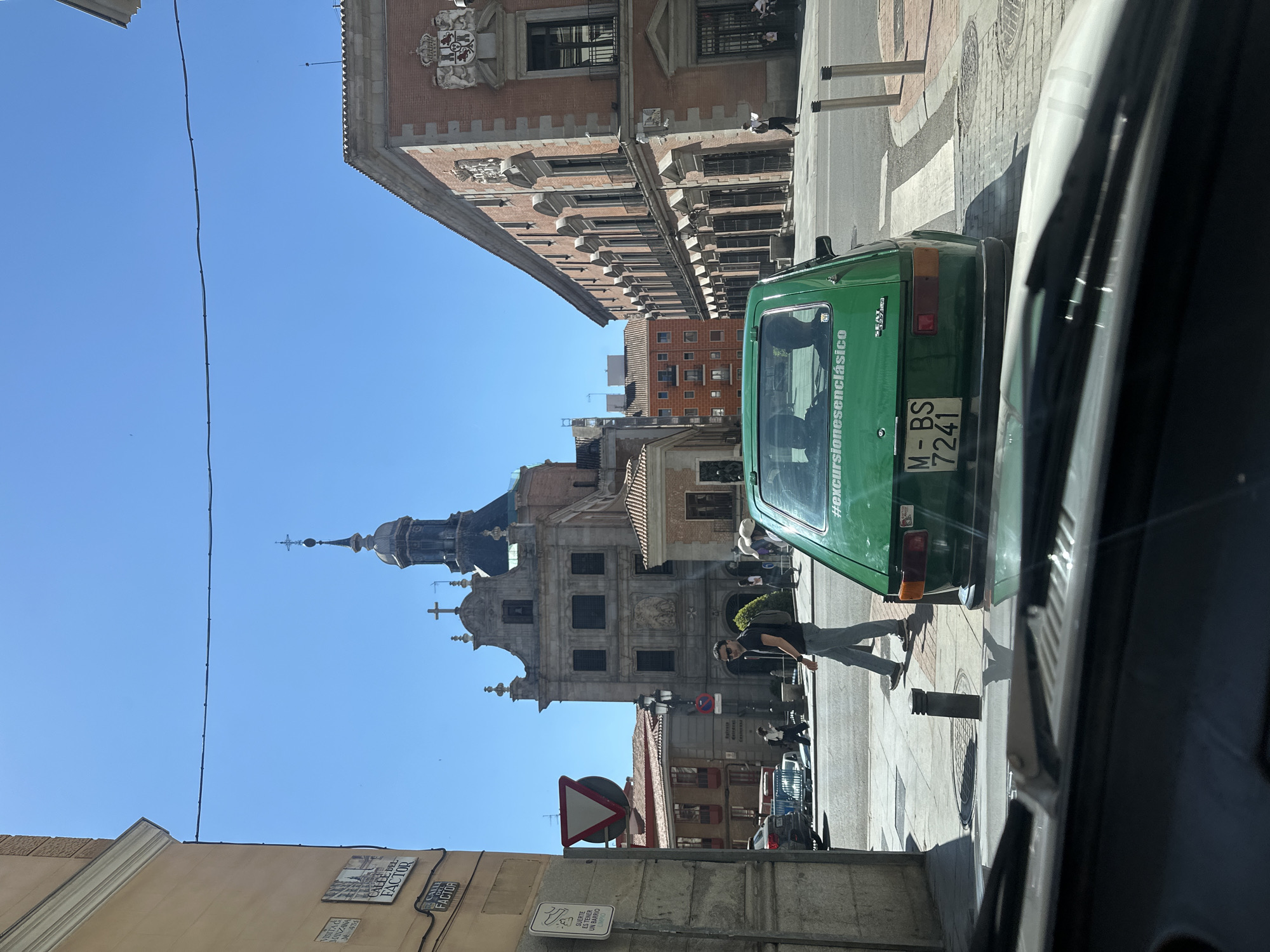 This image depicts a sunny street scene in Madrid, Spain, under a clear blue sky. In the foreground, a vibrant green classic car, possibly a Seat 600, is prominently featured. The car bears the text "#excursionesenclásico" on its side, indicating its use for classic car tours, and its Spanish license plate "M-BS 7241" confirms its Madrid registration.
A person wearing sunglasses and carrying a backpack is seen walking on the sidewalk next to the green car. In the background, a magnificent, ornate building with a distinctive dome and spire dominates the skyline, characteristic of historical churches or significant architectural landmarks in Madrid, such as the Real Colegiata de San Isidro. To the right of this building, another large, multi-story brick building with numerous windows and decorative cornices is visible.
Several street signs and plaques are present, with "CALLE DEL FACTOR" (Factor Street) clearly visible on a street sign. Another plaque reads "SUERTE ES TENER UN BARRIO LIMPIO" (It's lucky to have a clean neighborhood). The bright, direct sunlight creates strong shadows, suggesting it is midday or early afternoon. The overall scene conveys a pleasant, typical day in a historic European city, blending local life with elements of tourism through the classic car.