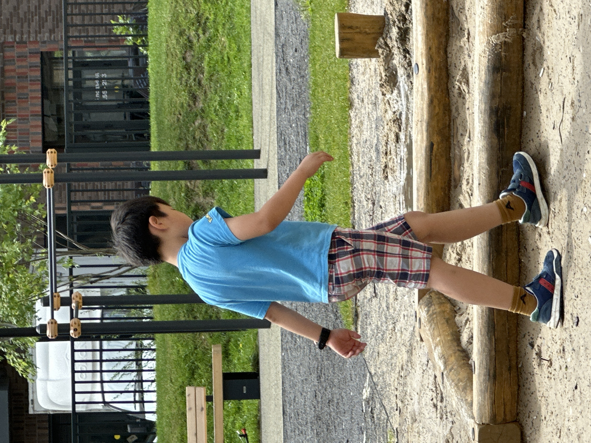 A young boy with dark hair, wearing a blue t-shirt and plaid shorts, walks across a sandy playground area in Znamya Oktyabrya, Russia. He is wearing dark sneakers with red accents and brown socks. A black fitness tracker is visible on his left wrist. The playground features wooden logs arranged as part of the play structure, and a small wooden post stands in the sand to the right. In the background, a metal fence surrounds a grassy area with a bench. Beyond the fence, a white truck and a brick building are partially visible. The environment appears to be an outdoor public space.