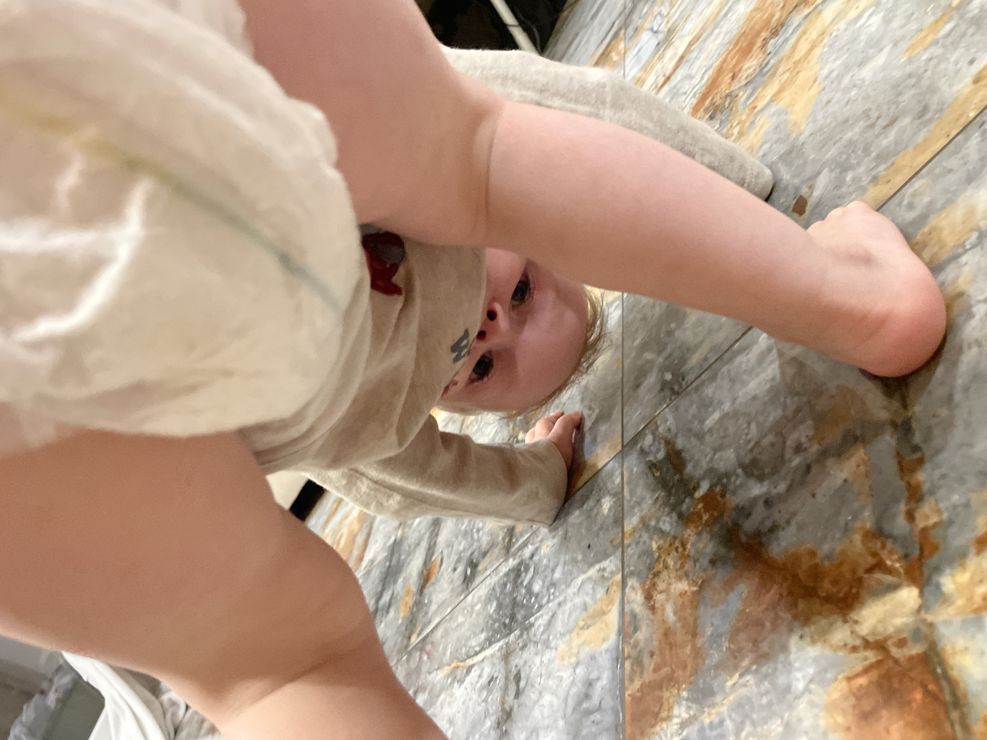 A close-up, low-angle shot shows a baby on a tiled floor. The baby, wearing a diaper and a beige long-sleeved shirt, is bent over with its legs visible in the foreground, obscuring much of the scene. The baby's face is visible as it looks towards the camera. The floor appears to be tiled with a marbled pattern in shades of gray, beige, and brown. The location is Unknown, Unknown.