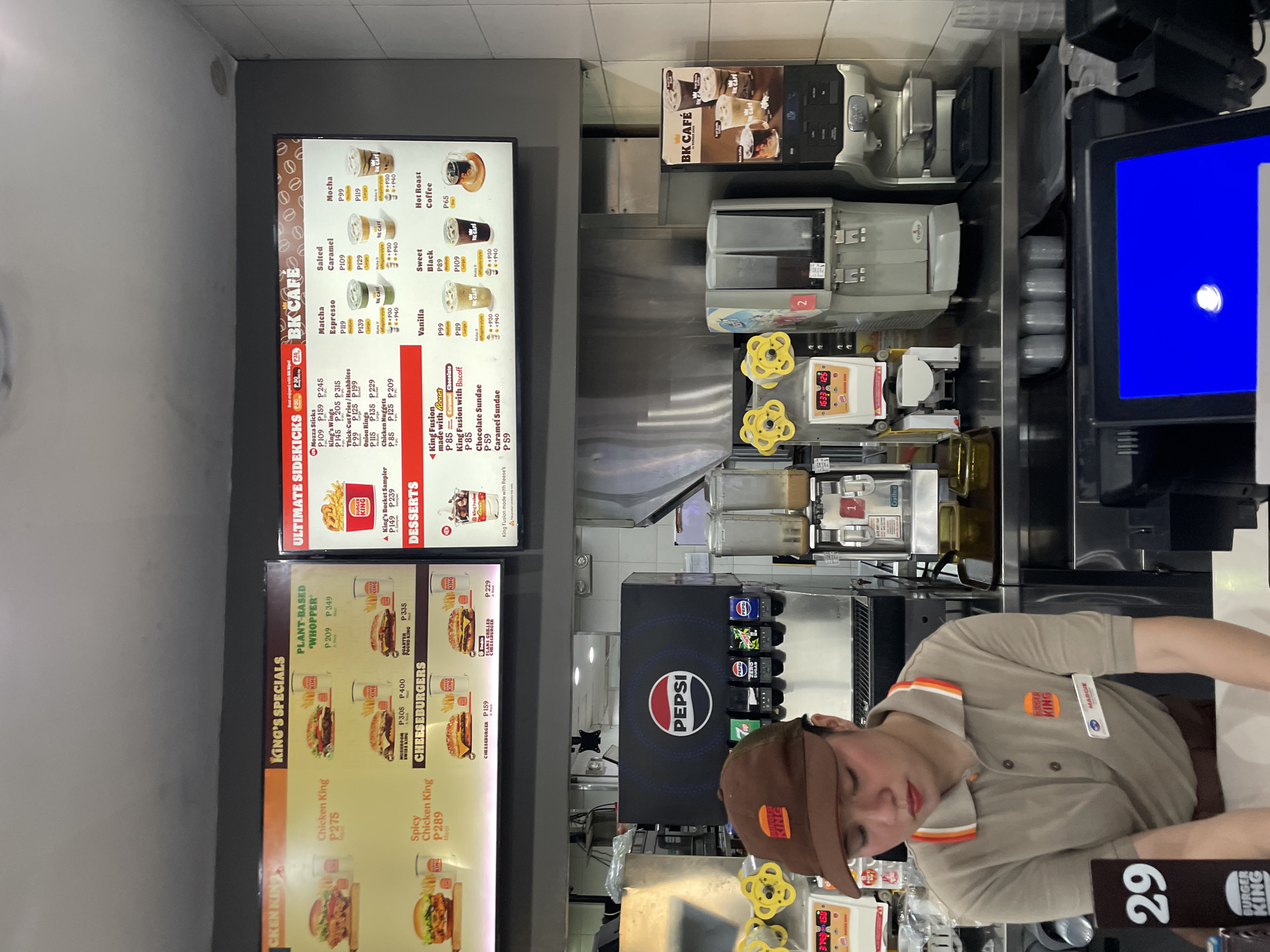 The image is a medium shot of a Burger King counter in Davao City, Philippines. A female employee wearing a brown Burger King uniform and cap is standing behind the counter. She is looking down and appears to be preparing an order. To her left are several drink dispensers, including a Pepsi dispenser. To her right are more drink dispensers and a menu board displaying food and drink items. The menu board is divided into sections such as "King's Specials," "Ultimate Sidekicks," "BK Cafe," and "Desserts." The overall environment appears to be a fast-food restaurant.