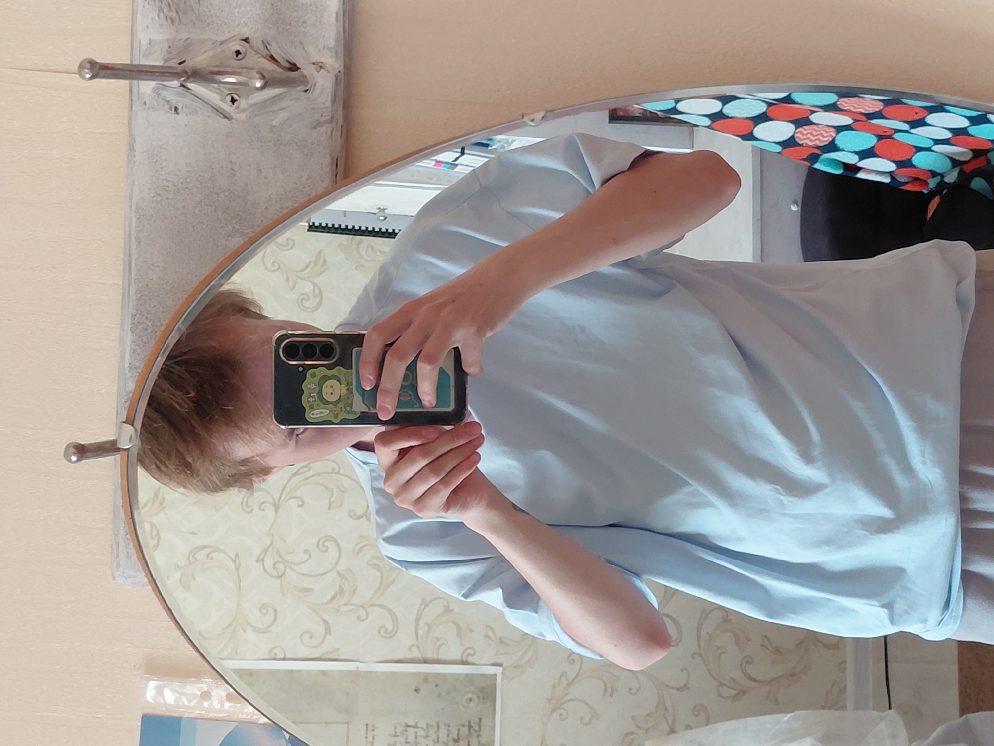 An individual is captured in a mirror selfie, with the image rotated 90 degrees counter-clockwise. The person, with short light brown hair, is wearing a light blue short-sleeved t-shirt and light-colored pants or shorts. They are holding a dark-colored smartphone with a light green/yellow cartoon-themed case, featuring an animal character, to take the photo. Their face is partially obscured by the phone.
The mirror, which is oval or rounded rectangular with a light wooden frame, reflects an indoor setting, likely in Nadym, Russia. The background features a wall covered in light-colored wallpaper with a subtle, swirling floral pattern. To the upper left of the mirror, a silver, two-pronged clothes hook is mounted on a textured, light grey wall panel. In the reflection, within the upper right curve of the mirror, a vibrant fabric with a pattern of red, white, and teal polka dots or circles is visible. Below the mirror, some white papers or plastic sheets with unreadable text or graphics are present. The overall scene depicts a casual indoor moment.