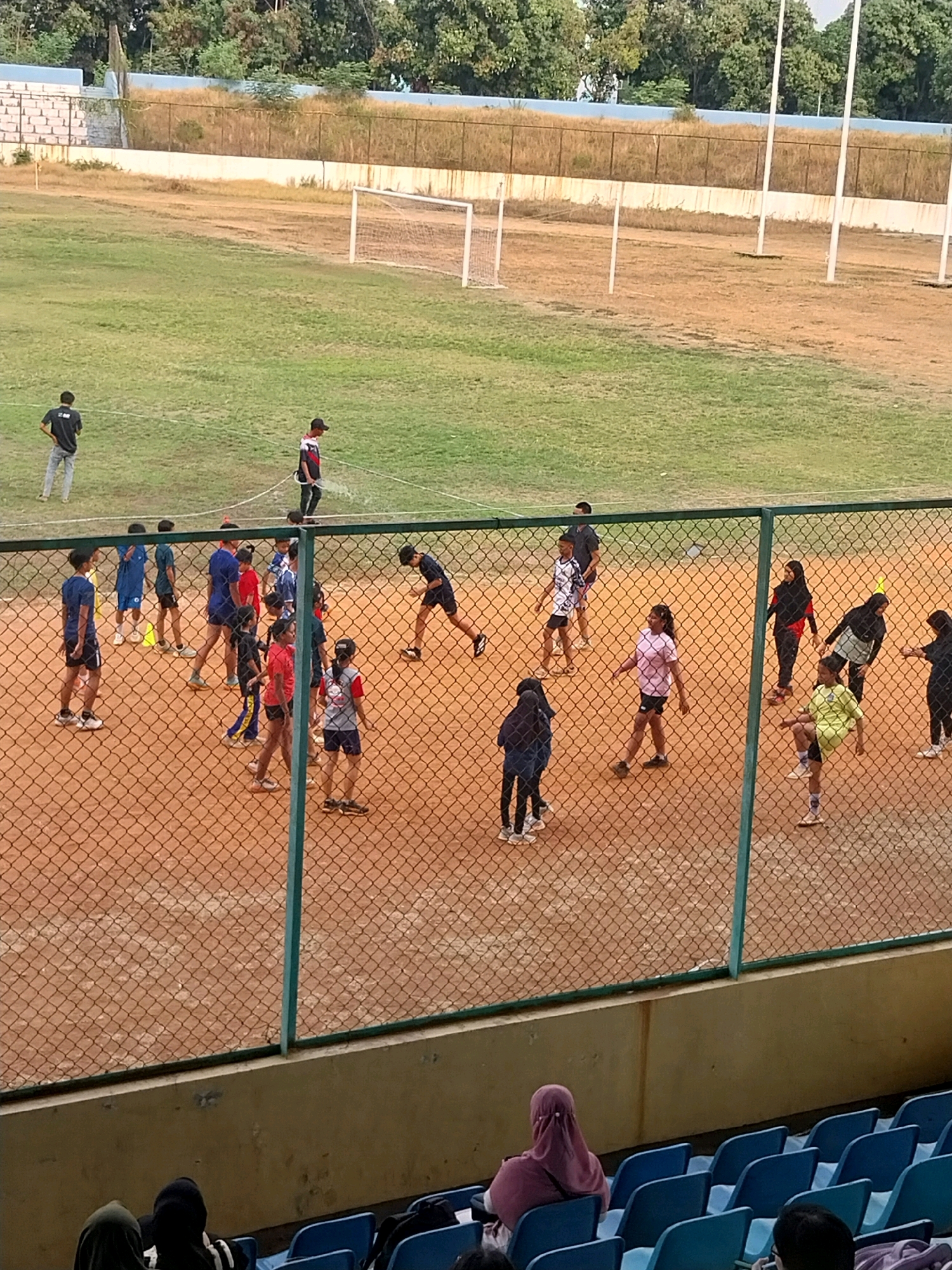 The image depicts a group of individuals, predominantly children and teenagers, engaged in athletic activities on a dirt field within a stadium. The field appears to be a multi-purpose sports area, with a grassy section in the background that suggests it might be a football field, evidenced by a goalpost visible in the upper left corner. The foreground is dominated by a fenced-off dirt area where the participants are gathered. A chain-link fence with green metal posts separates the playing field from a seating area in the lower portion of the image, where some spectators are visible. The stadium itself seems to be located outdoors, with trees and a fence line visible in the background, indicative of an urban or suburban setting. The time of day appears to be late afternoon, given the lighting conditions and the long shadows cast by some individuals. The overall atmosphere suggests a training session or a casual sports event. The presence of diverse attire, including athletic wear and headscarves on some individuals, points to a varied group of participants. The scene is characteristic of a local sports facility, possibly within the city of Cirebon, Indonesia, where such outdoor stadiums are common for community sports and recreational activities.