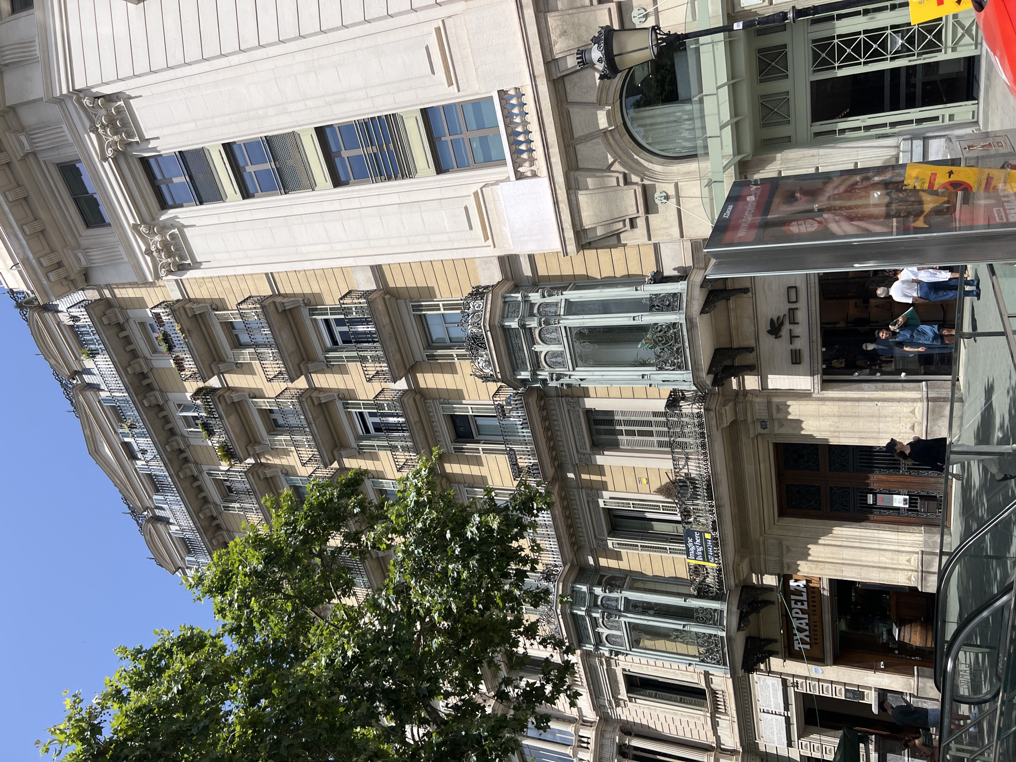 The image captures a street view in Barcelona, Spain, focusing on the architecture of a building with a pale yellow facade. The building features multiple floors with numerous balconies adorned with ornate black railings. Some balconies have plants, while others are empty. Several windows have dark shutters, some open and some closed. A distinctive feature is a bay window on a lower floor, elaborately decorated with green metalwork and decorative glass panels. To the right, a contrasting building with a lighter, creamy facade displays neoclassical architectural elements, including pilasters and decorative cornices.
At street level, two storefronts are visible. One displays the signage "TXAPELA," indicating a food establishment. Next to it is a storefront with the "ETRO" logo. Several people are present on the sidewalk. A person in a black top and light-colored pants walks towards the right. Another individual, a man wearing a dark shirt and shorts, stands near the "TXAPELA" entrance, looking at a phone. Further to the right, a large advertising display with an image of a person wearing yellow clothing stands prominently. A lamppost with a dark, ornate fixture is situated near the right edge of the frame. Trees with lush green foliage partially obscure the upper floors of the building on the left. A red car is partially visible in the lower right corner. The sky is a clear, bright blue.