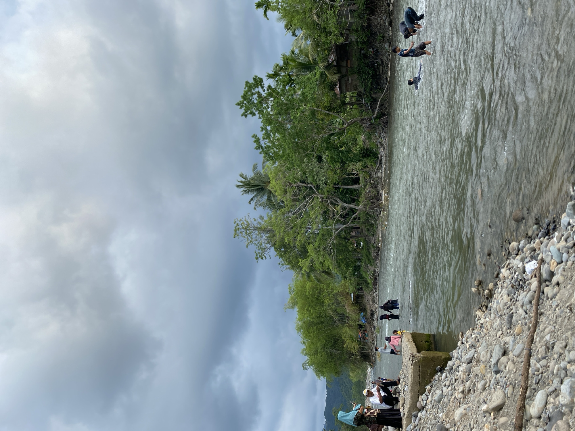 The image depicts a river scene in Meurah, Indonesia, under a cloudy sky. A group of people is visible, with some individuals swimming or wading in the river's shallow waters. Along the riverbank, lush vegetation including trees and bamboo thrives. On one side, a stone embankment or structure is present, with people gathered nearby. The riverbed appears rocky.