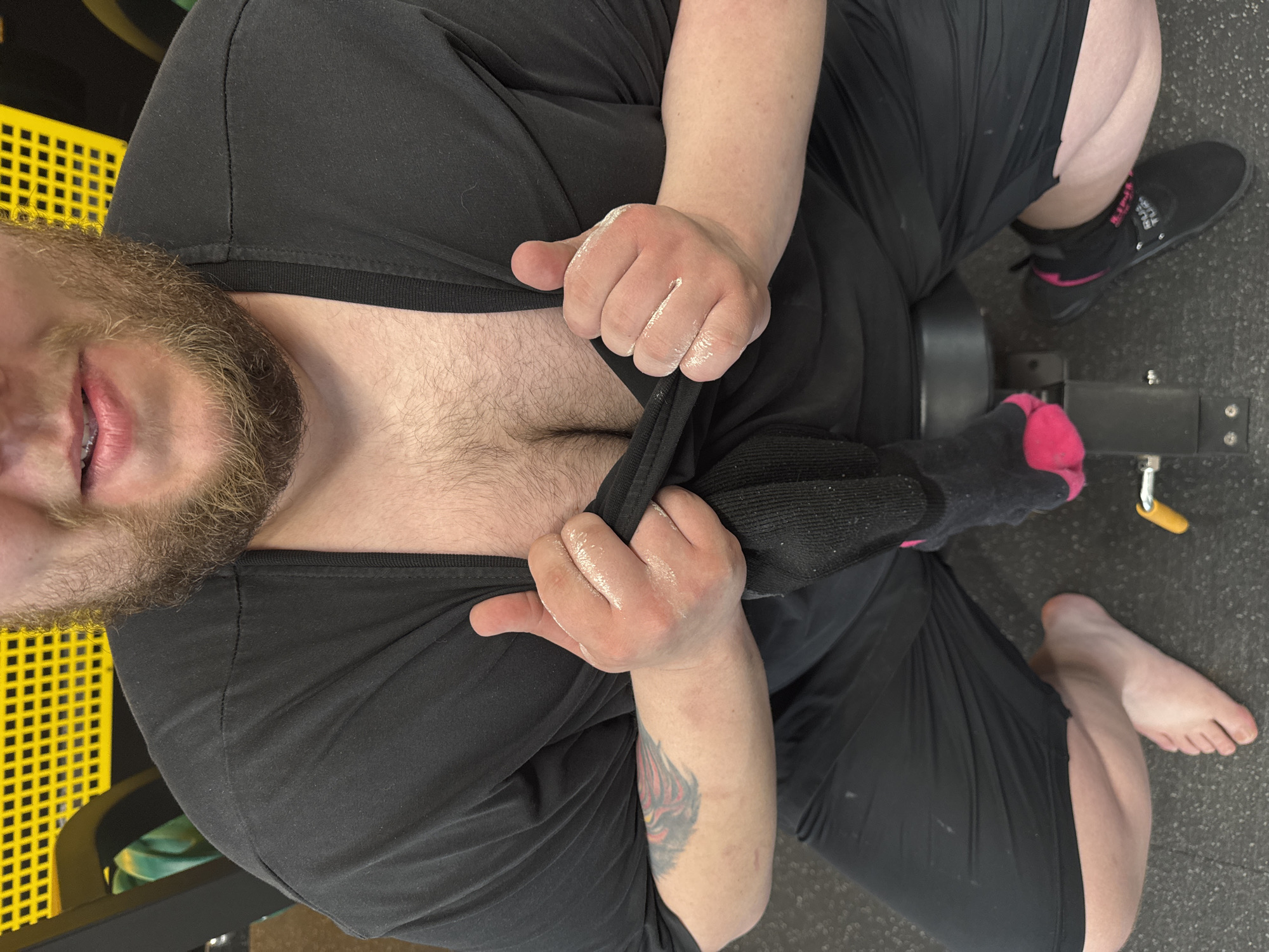 A medium close-up, high-angle shot captures an individual from the chest down, with the lower portion of their face, including a light brown beard and a slight smile, visible. The individual has light skin and a substantial amount of brown chest hair. They are wearing a black short-sleeved t-shirt and black shorts, and are seated on a dark gray padded gym bench. Both hands, which appear slightly reddened and are covered in a white, chalk-like substance, are gripping and pulling the t-shirt open at the neckline, exposing the upper chest area. A tattoo with red, orange, and yellow elements is visible on the inner portion of the left forearm. The individual's left foot is bare and rests flat on a dark gray, speckled gym floor. The right foot, positioned near a dark metallic structure with a yellow handle, is wearing a black athletic shoe with pink accents and a black sock with a pink toe area. The background features a bright yellow, grid-patterned surface, consistent with gym equipment or a wall, along with dark structural elements. The scene is an indoor gym in Unknown, Unknown.
