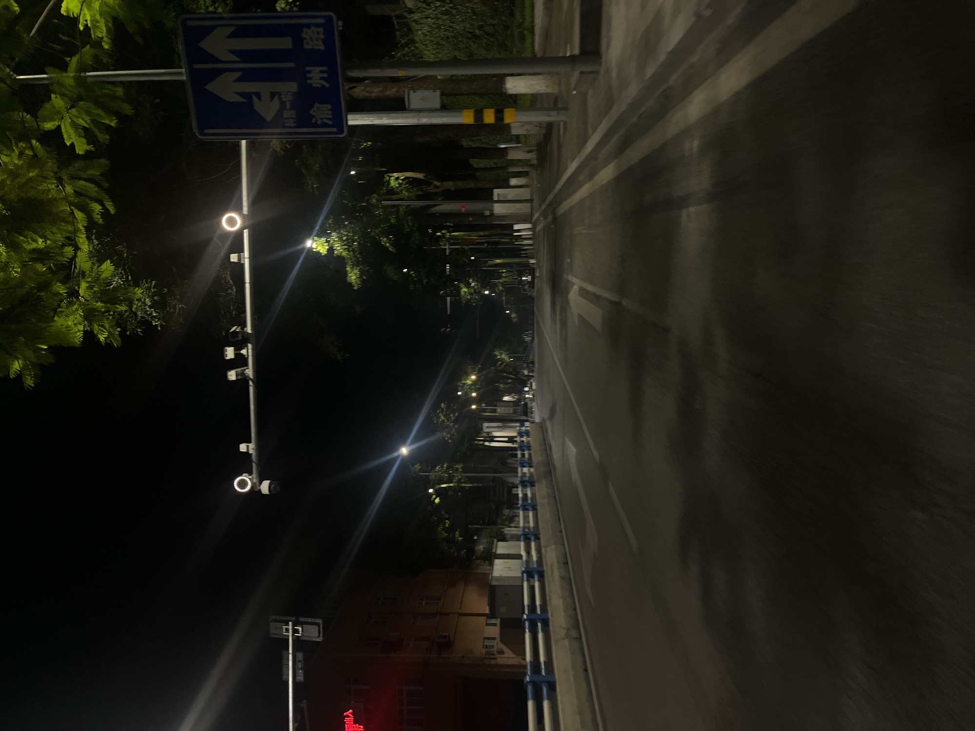 This image captures an empty urban road at night, identified as Yuzhou Road (渝州路) near Keyuan 1st Street (科园一街) in Jiulongpo District, Chongqing, China. The road surface appears dark and reflective, suggesting dampness, possibly from rain. The perspective is low, likely taken from a moving vehicle.
In the midground, a concrete median barrier with blue and white railings separates the main thoroughfare from the left side of the road. To the left, a multi-story building displays a red illuminated sign or interior light. Above the road, an overhead gantry spans the width, equipped with a prominent circular light (possibly a traffic sensor or signal) and multiple surveillance cameras or sensors. A vertical traffic light is visible further to the left.
On the right side of the road, a blue rectangular directional sign is affixed to a pole, clearly displaying "渝州路" (Yuzhou Road) in large characters, accompanied by arrows indicating straight and left turns. Smaller text below reads "科园一街" (Keyuan 1st Street). Lush green trees with dense foliage overhang the road from the right, extending into the distance along a sidewalk. Streetlights cast bright glares and lens flares across the scene, providing the primary illumination. The background shows the road receding, lined by more trees and indistinct building outlines. No people or active vehicles are visible, contributing to the deserted nighttime atmosphere.