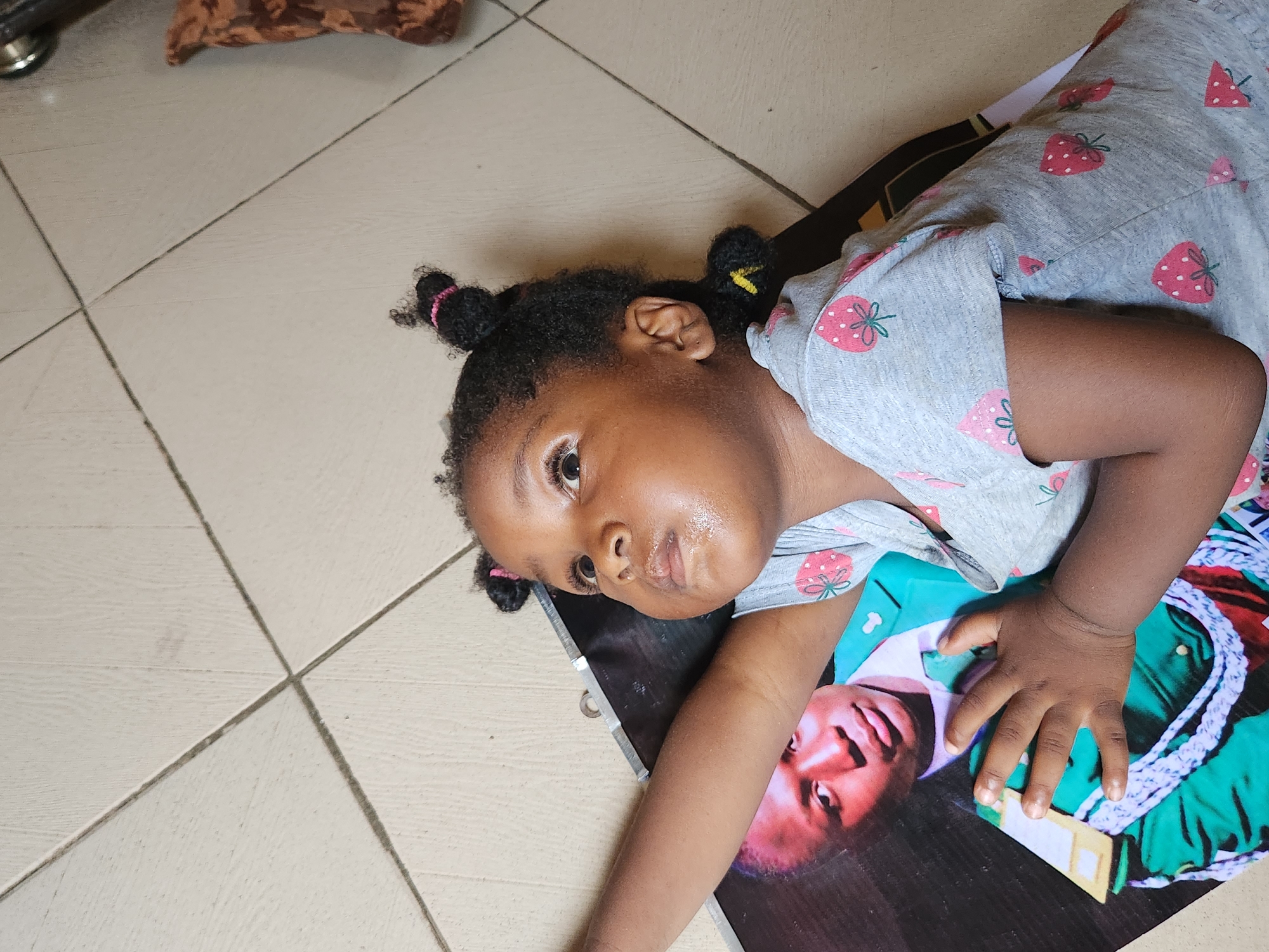 A young Black girl with dark curly hair pulled into two top buns and a side ponytail is lying on a tiled floor. She is wearing a grey t-shirt adorned with red strawberry patterns. Her left arm is extended, and her hand rests on a large printed photograph of a Black man in a green military uniform with white trim and medals. The man in the photograph has dark skin and is looking towards the left of the frame. The floor is composed of light-colored square tiles with visible grout lines. To the right of the girl, a portion of a patterned brown textile object is visible. The lighting suggests an indoor setting, potentially in Kaduna South, Nigeria, given the context.