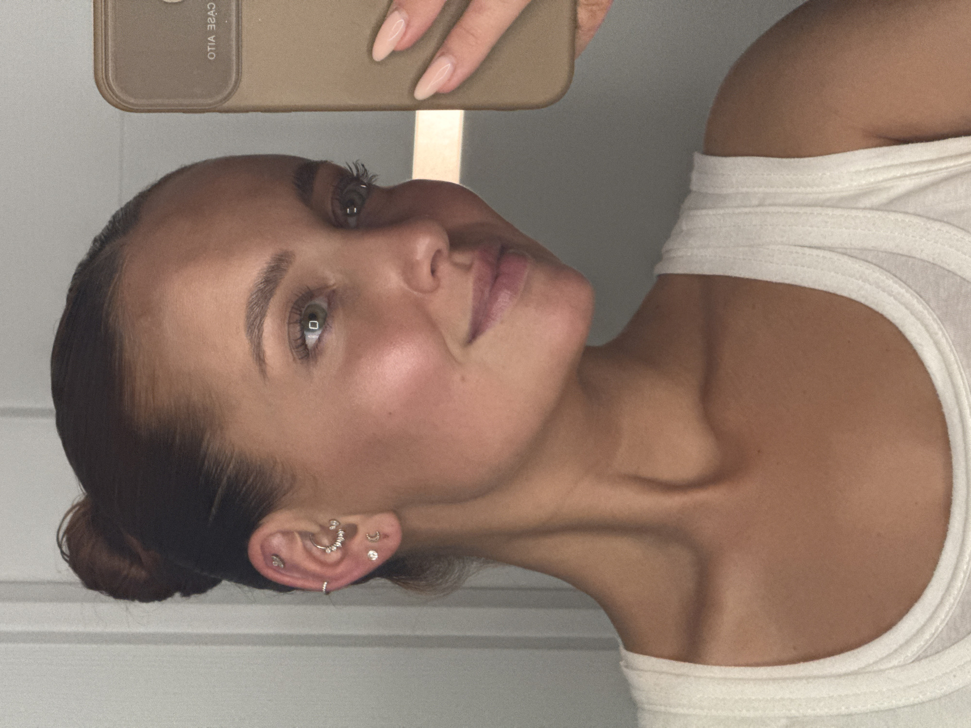 The image is a close-up selfie of a young Caucasian woman with her hair pulled back in a bun. She is wearing a white tank top and several earrings in her left ear, including a hoop and stud earrings. She is holding a tan smartphone in her right hand, which is partially visible. The background appears to be indoors, with a wall that has vertical lines. There is no indication of the location being in Chaikovsky, Russia.