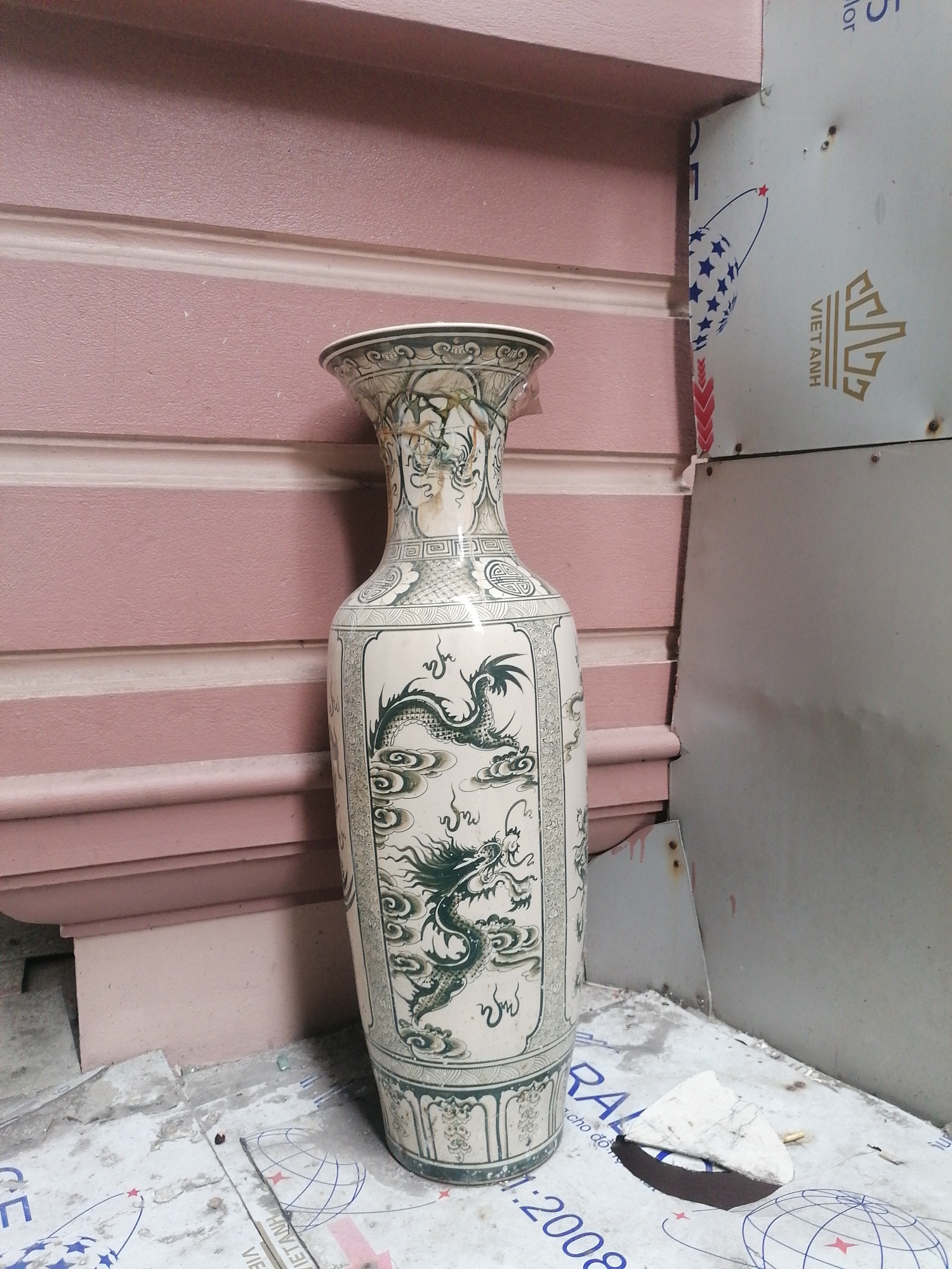 A tall, ornately decorated ceramic vase stands on a surface with printed designs in what appears to be an urban or semi-industrial setting. The vase is predominantly cream-colored with green dragon motifs and intricate geometric patterns. It has a narrow neck that widens at the top and a broader base.
The backdrop includes a pink exterior wall with layered molding and a stack of metal panels, one of which has a graphic design possibly related to a Vietnamese brand. The floor or surface where the vase sits is covered with printed material featuring text and a globe design. A small piece of white debris lies nearby.
The location is suggested to be Bai Chay Ward, Vietnam. There are no people visible in the image, focusing on the artifact and its immediate surroundings.