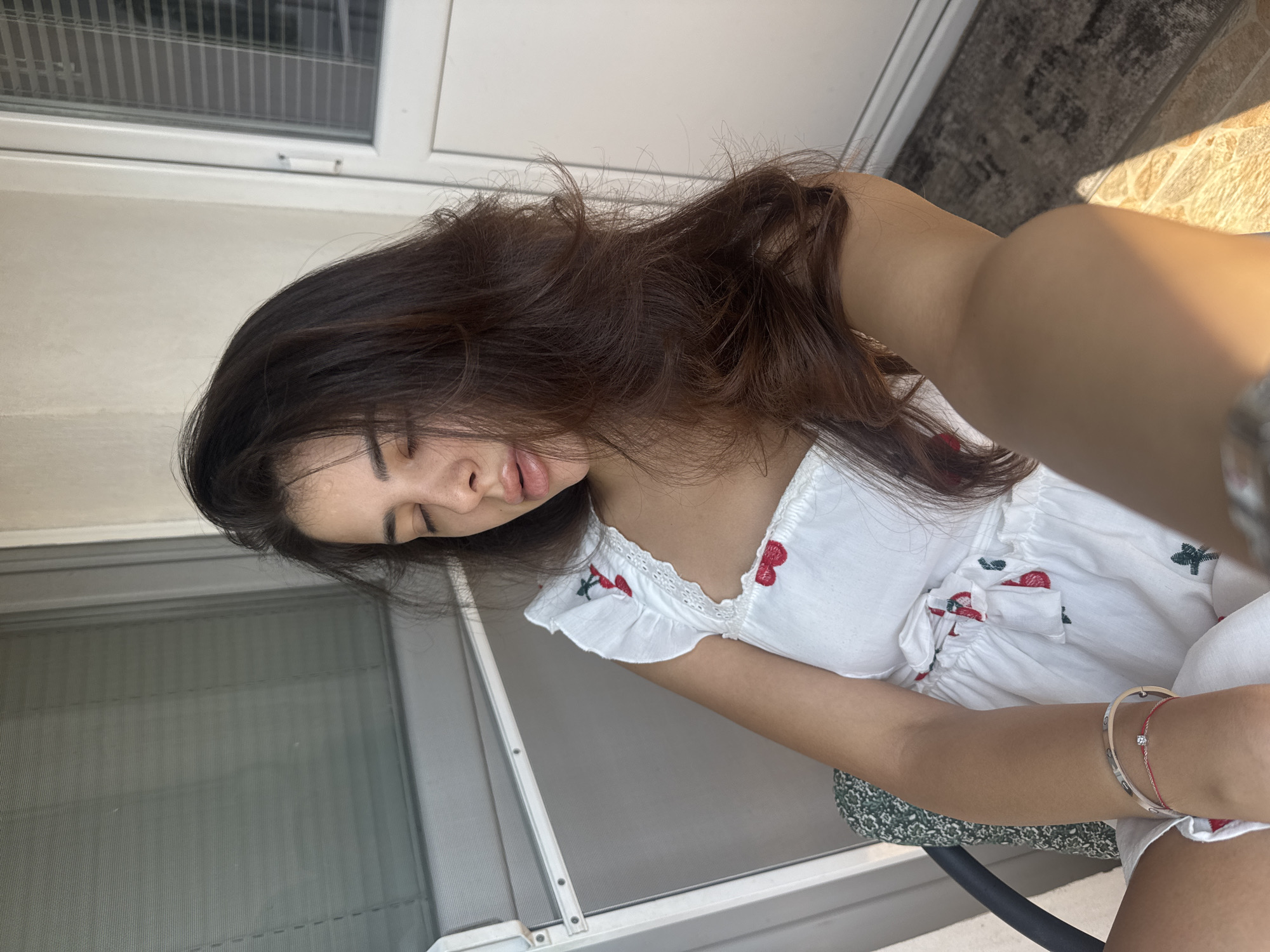 A woman is sitting in a chair, possibly on a balcony, with her eyes closed and her head tilted back. She has long, dark brown hair that falls around her shoulders. She is wearing a white dress with a red cherry pattern and ruffles around the neckline. There is a bracelet on her left wrist. The background shows a window with blinds and a white door. The lighting suggests it is daytime. The location is not specified.
