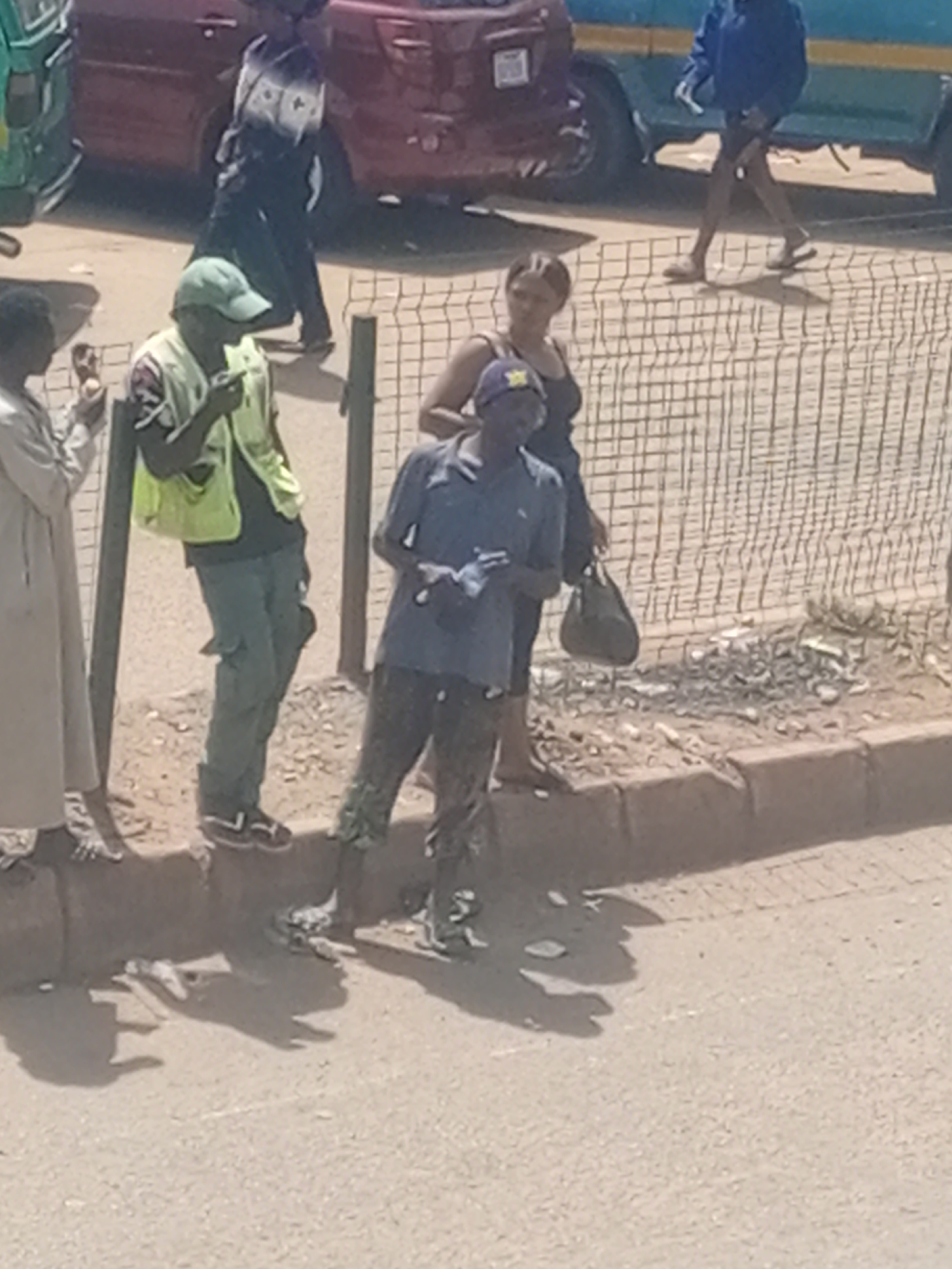 In a medium shot, three men and one woman are visible on a dirt road next to a curb. A green three-wheeled vehicle and a red SUV are partially visible in the background. A light blue vehicle with yellow accents is also in the background. A wire fence runs along the curb behind the people.
The person on the far left is wearing a long, light-colored robe. Next to them, a man in a bright yellow vest and a green baseball cap is holding something up to his mouth. To the right of him, a man in a blue polo shirt and a blue baseball cap with a yellow emblem is standing with his hands in front of him. Behind him, a woman in a black tank top is looking to her right.
The men appear to be interacting with each other. The man in the yellow vest has his mouth open and seems to be talking or eating. The man in the blue polo shirt is holding a white object in his hand. The woman is looking off-camera.
The scene appears to be outdoors during the daytime, with the sun casting strong shadows. The overall atmosphere is casual, with people gathered on the side of the road. The presence of vehicles and the general street scene suggest an urban or semi-urban environment. There are no visible signs or text in the image.
