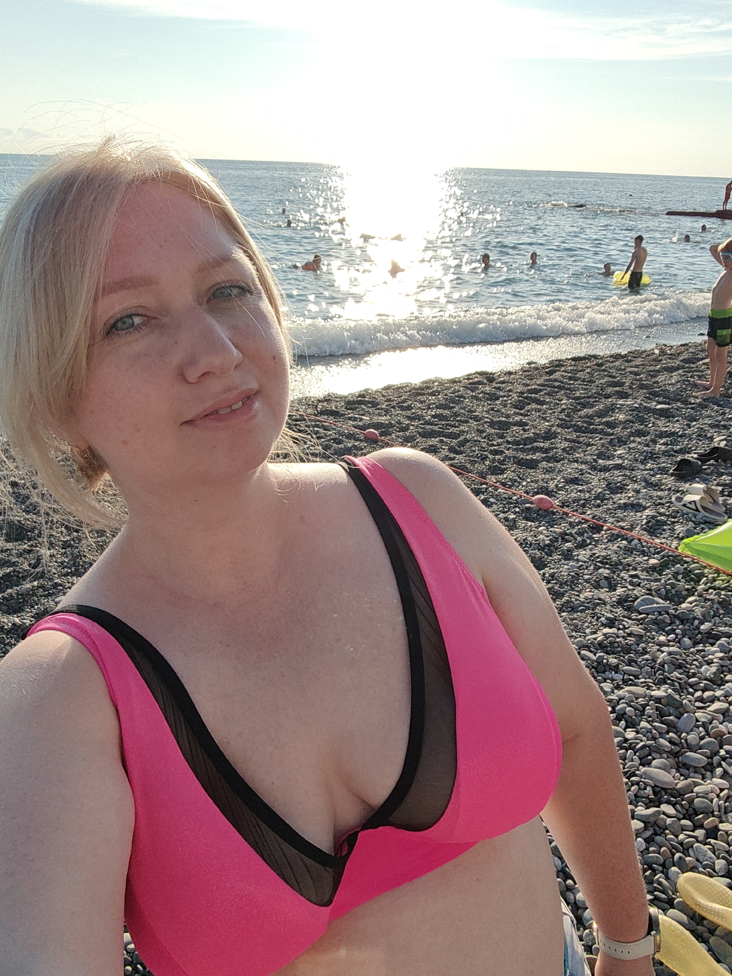 A woman with blonde hair and blue eyes takes a selfie on a sunny day at a pebble beach. She is wearing a bright pink bikini with black mesh accents. The ocean is in the background with several people swimming and playing in the water. The sun is shining brightly, creating a glare on the water's surface. The beach is covered in grey pebbles, and there are a few pairs of sandals and a yellow inflatable ring visible. The overall mood is relaxed and cheerful, suggesting a vacation or leisure day. The time of day appears to be late afternoon or early evening, given the golden light and the sun's position. The location is identified as Sochi, Russia. There is no visible text in the image.